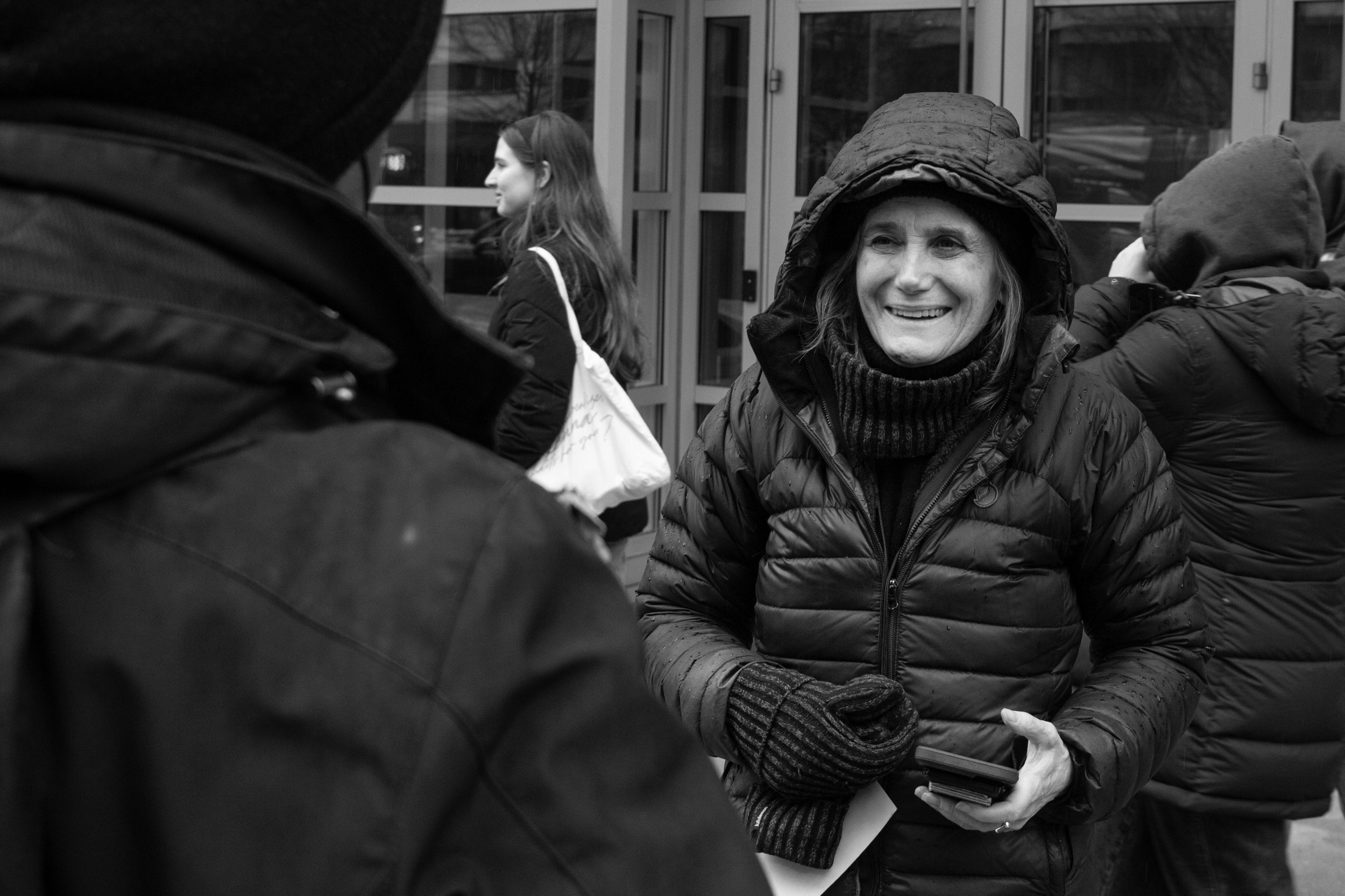 A woman smiling and wearing a hooded jacket, engaged in a conversation with other people outside a building.