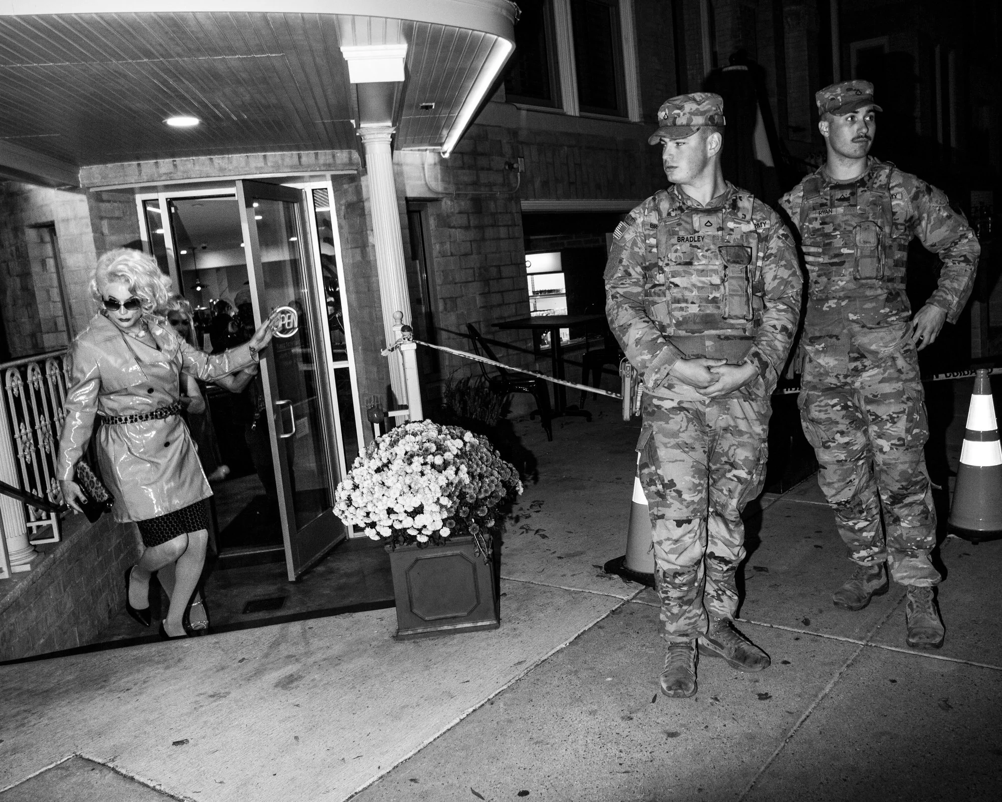 Two soldiers in camouflage uniforms standing outside a building at night, with a woman in sunglasses and a shiny coat near the entrance, holding a door handle, and flowers in a pot nearby.