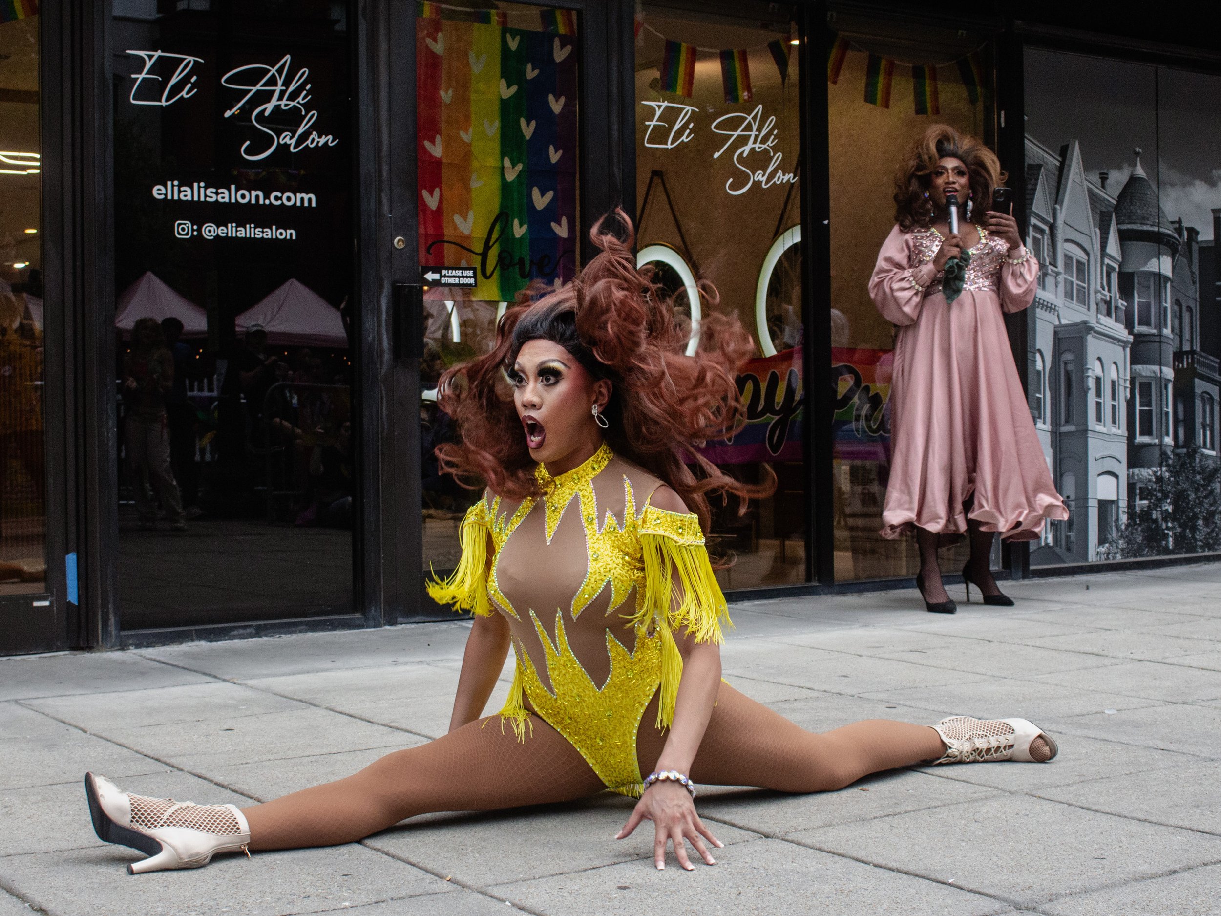 A drag performer in a bright yellow costume performing a split on the sidewalk with one hand on the ground, outside a salon decorated with rainbow pride flags and a rainbow pride flag poster. The performer has long, voluminous hair and dramatic makeu