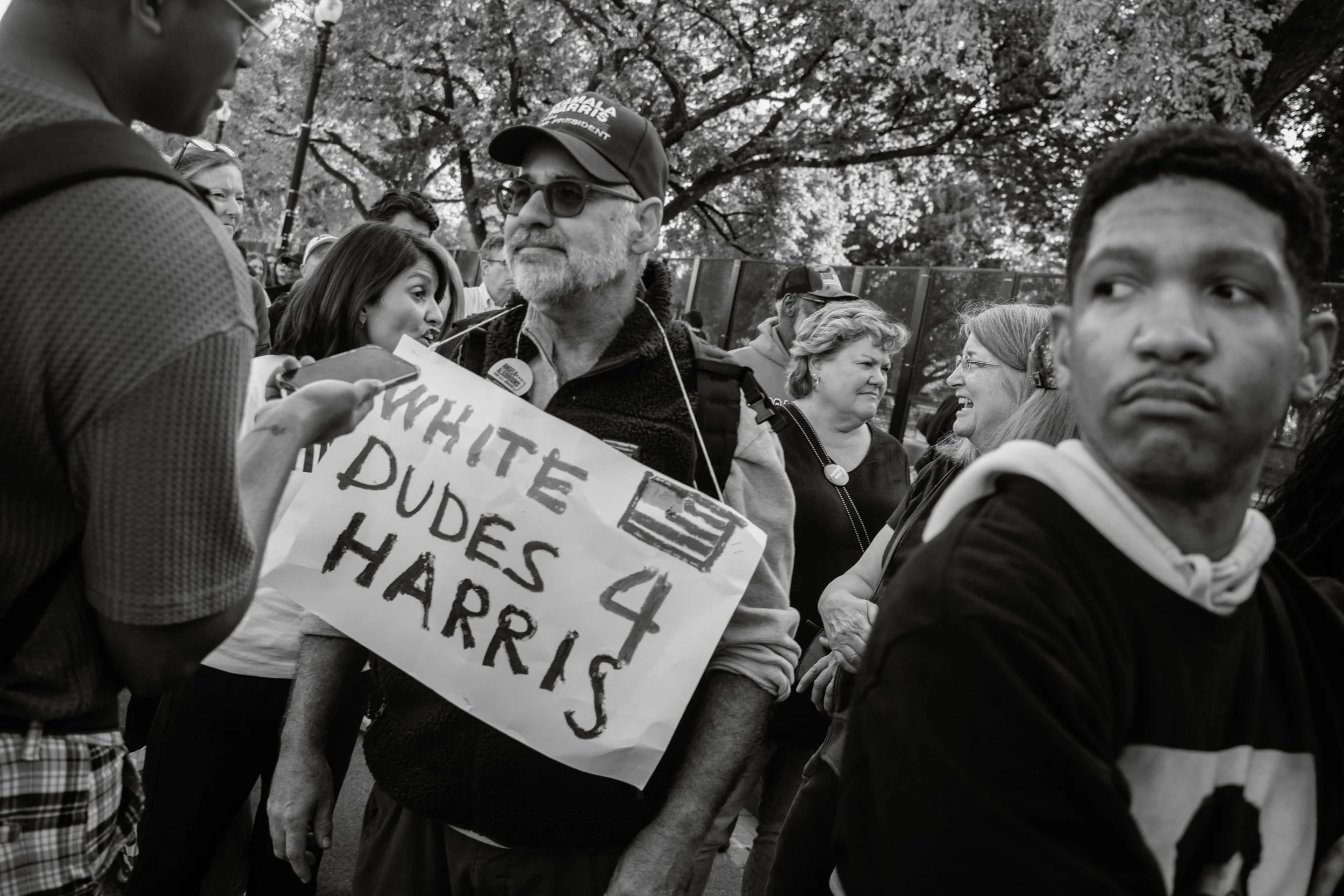 Black and white photo of a group of people gathered outdoors, some holding signs, involved in a protest or demonstration. Trees are visible in the background.