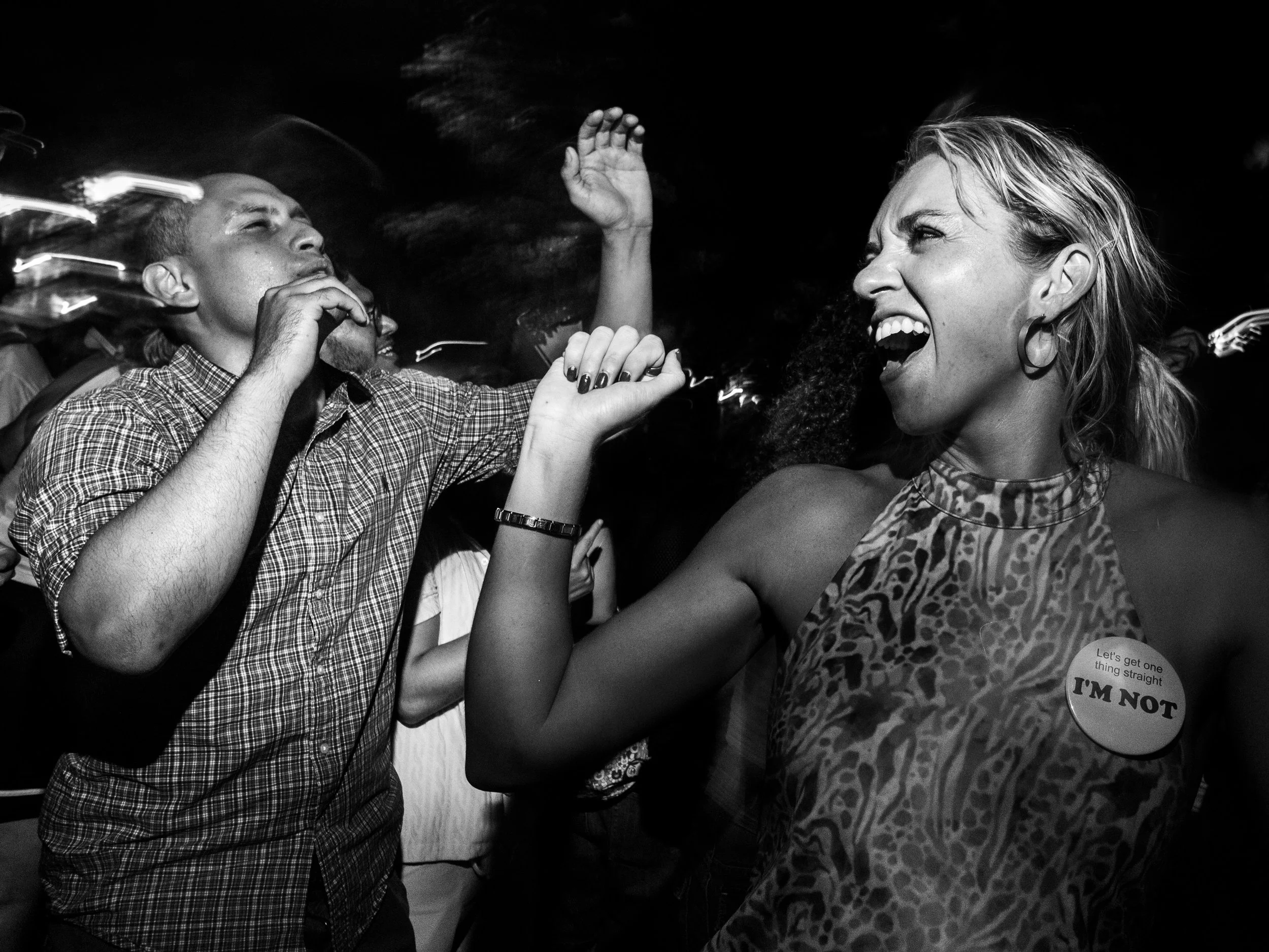 People at a social gathering, two women engaging in a playful arm wrestling match, one wearing a sticker that says "I'M NOT" and the other with her hair pulled back, both laughing.