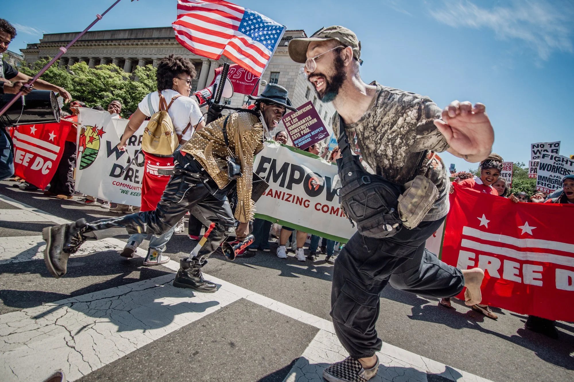 Protesters holding banners and flags, including an American flag with stars and stripes, march on a city street. The background features historic buildings and bright blue sky.