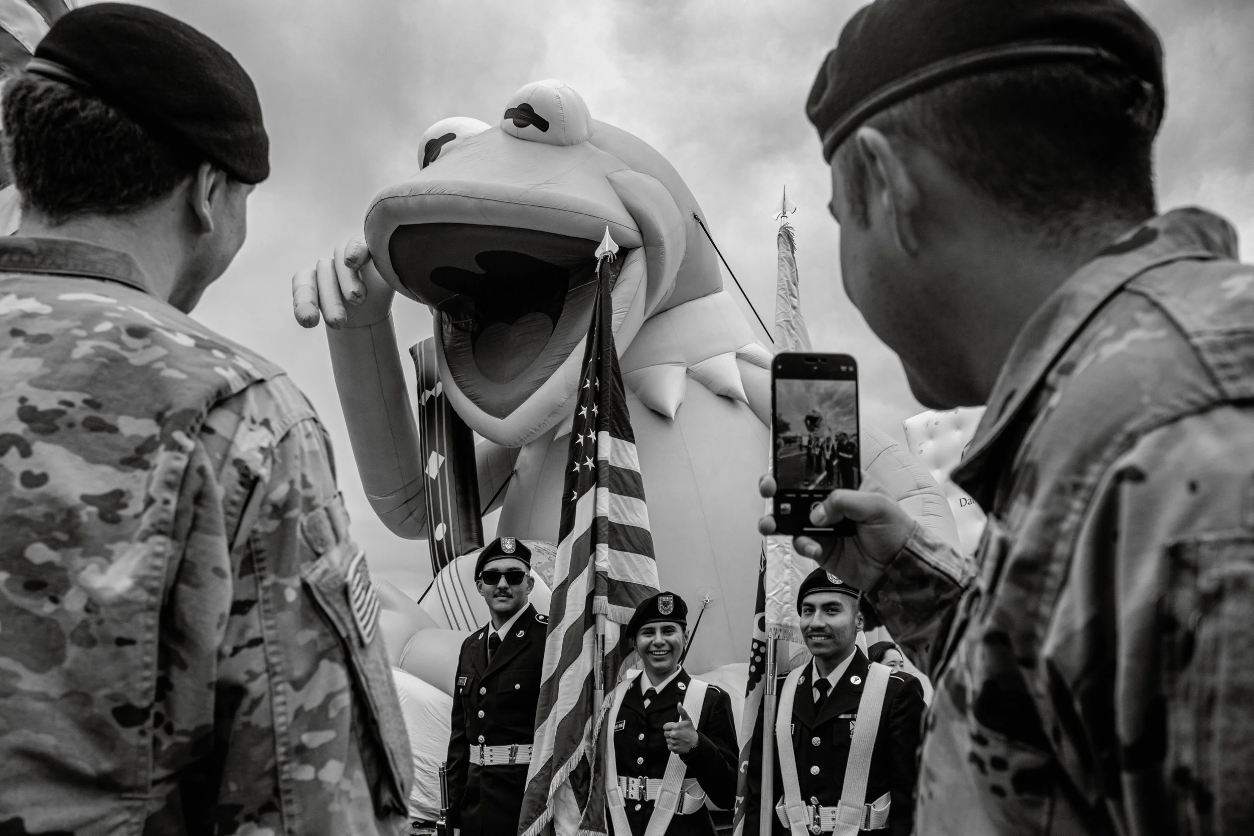 Black and white photo of people in military uniforms standing in a circle, with a large inflatable frog mascot holding flags in the background, during a parade or public event.