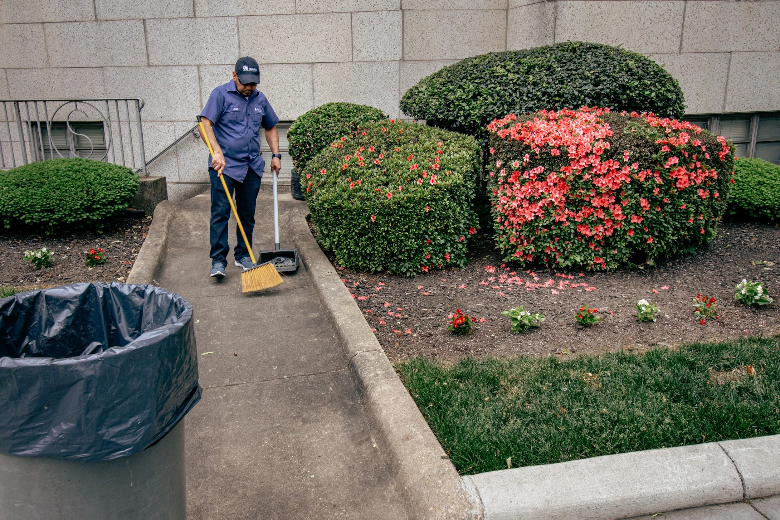 A man in a blue uniform raking leaves or debris in a small landscaped area with bushes and flowers, near a concrete wall, with a large trash can in the foreground.