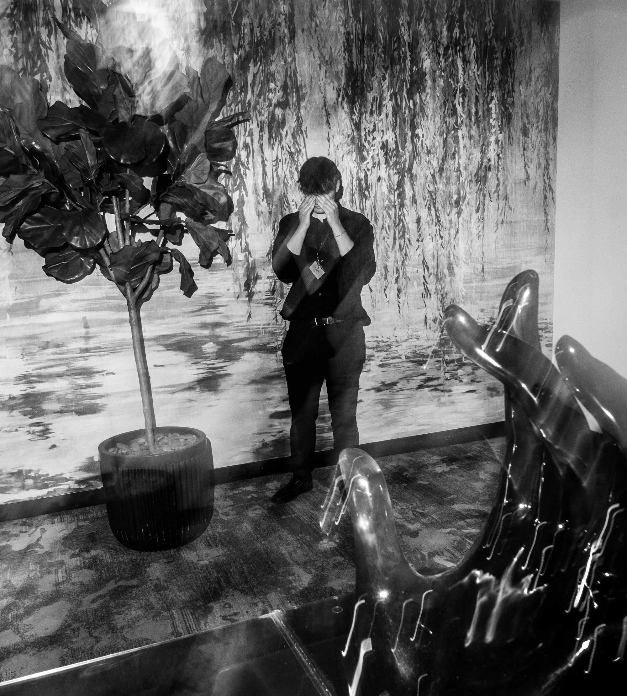 A person in dark clothing and a face mask is crouching and covering their face with their hands in front of a large painting, with a potted plant to the left and empty chairs in the foreground.