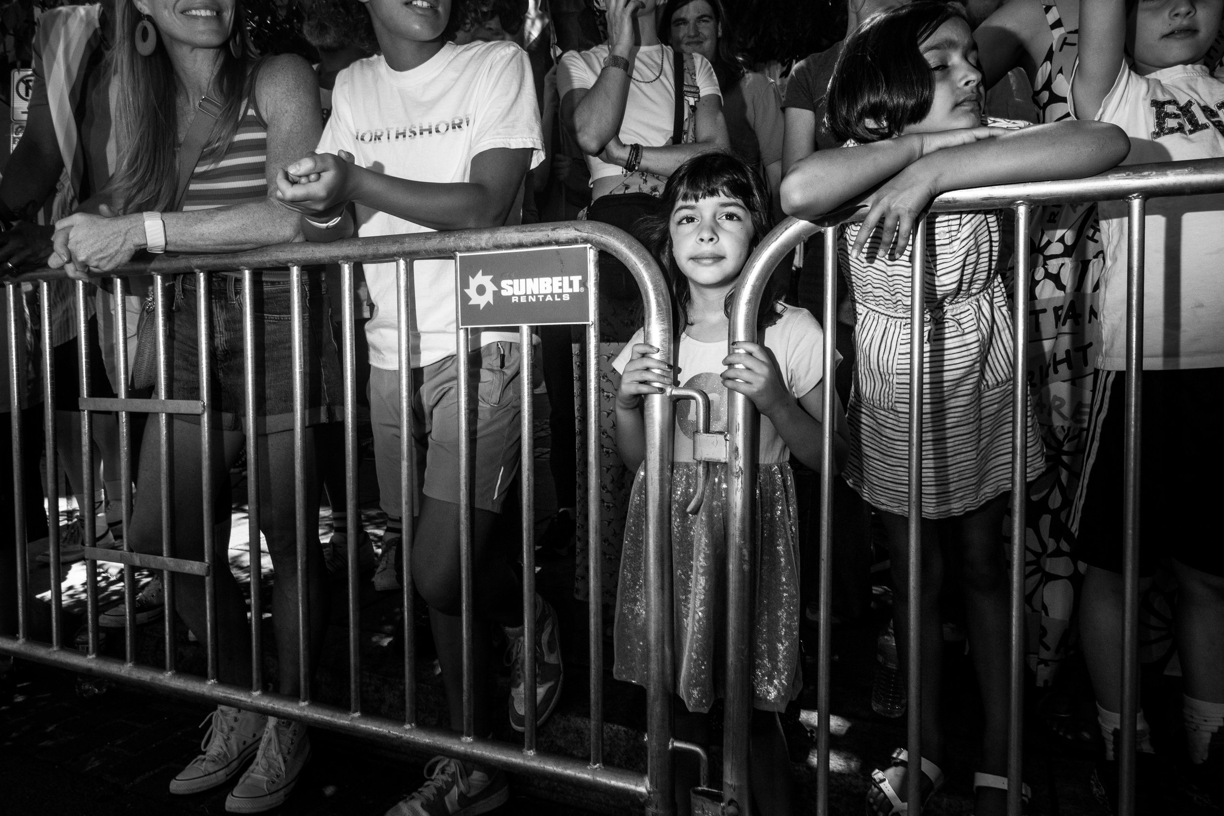 A group of children and adults standing behind a metal barricade at an event, with some children leaning on it and one girl holding onto the bars. The scene is in black and white.
