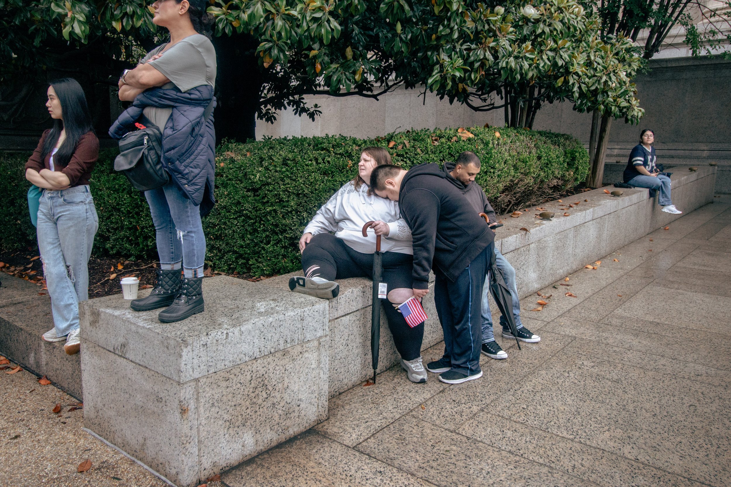 Several people sitting and standing on a concrete ledge outdoors. Some are looking at their phones or resting, with a large bush and trees in the background.