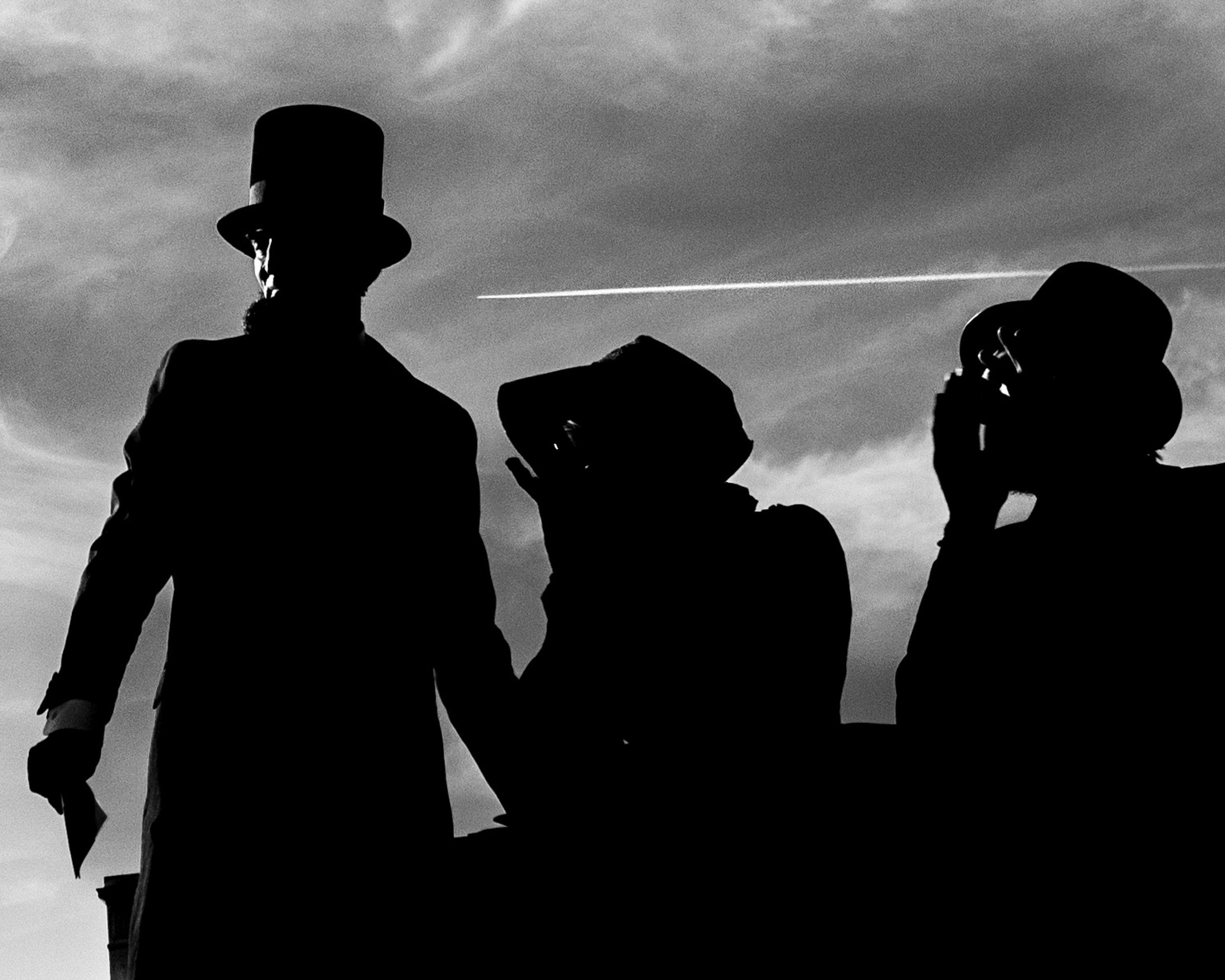 Silhouettes of three men dressed in vintage clothing, one wearing a top hat, against a cloudy sky with an airplane contrail.