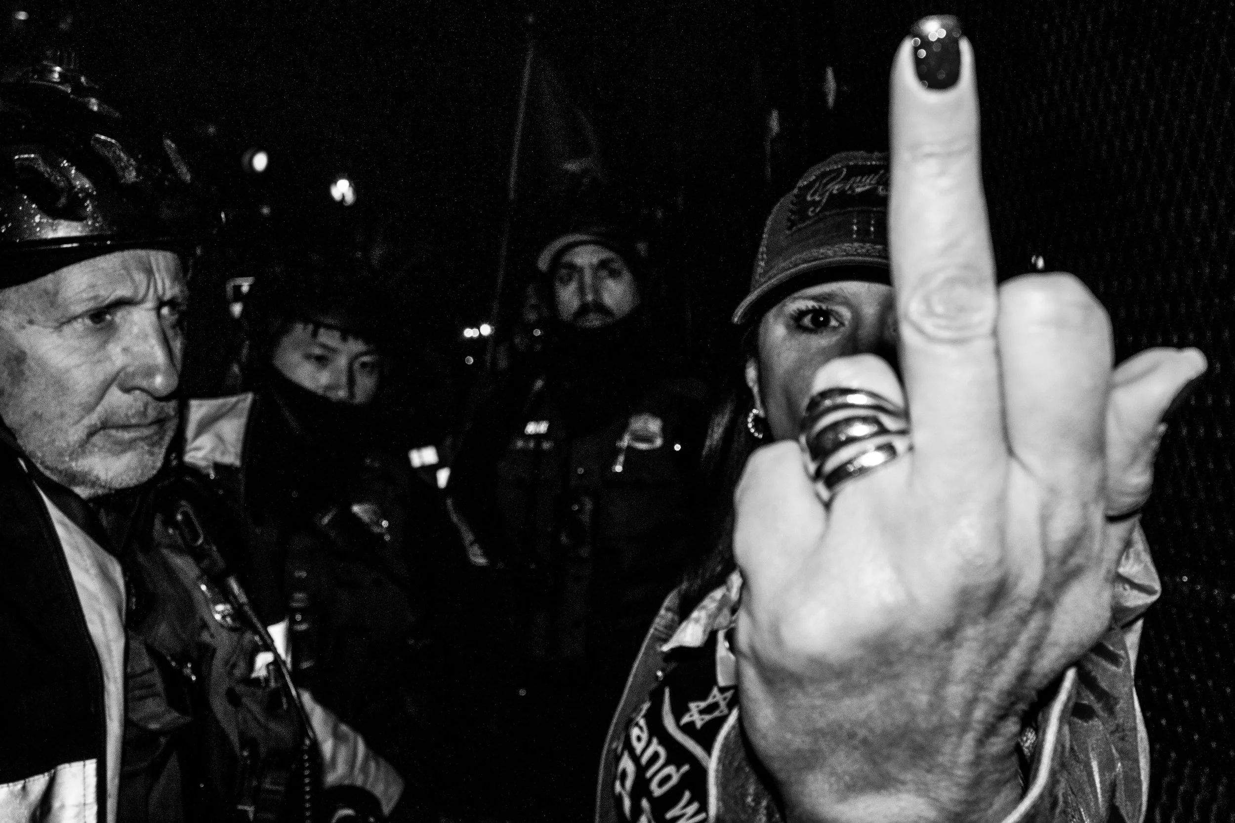 A black-and-white photo of a group of people, focusing on a woman showing her middle finger. The woman wears a beanie and rings on her fingers, with some text on her sleeves and a patch on her jacket. Other people in the background wear jackets and h
