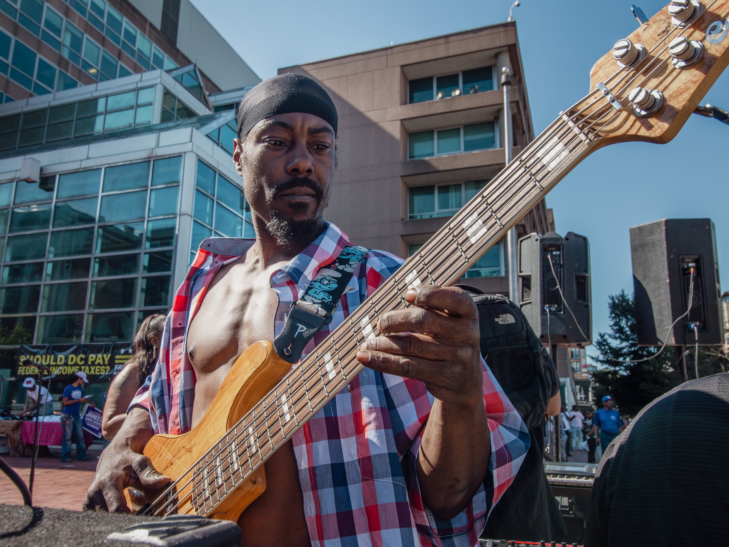 A man playing an electric bass guitar outdoors during a public event or protest, with buildings and people in the background.