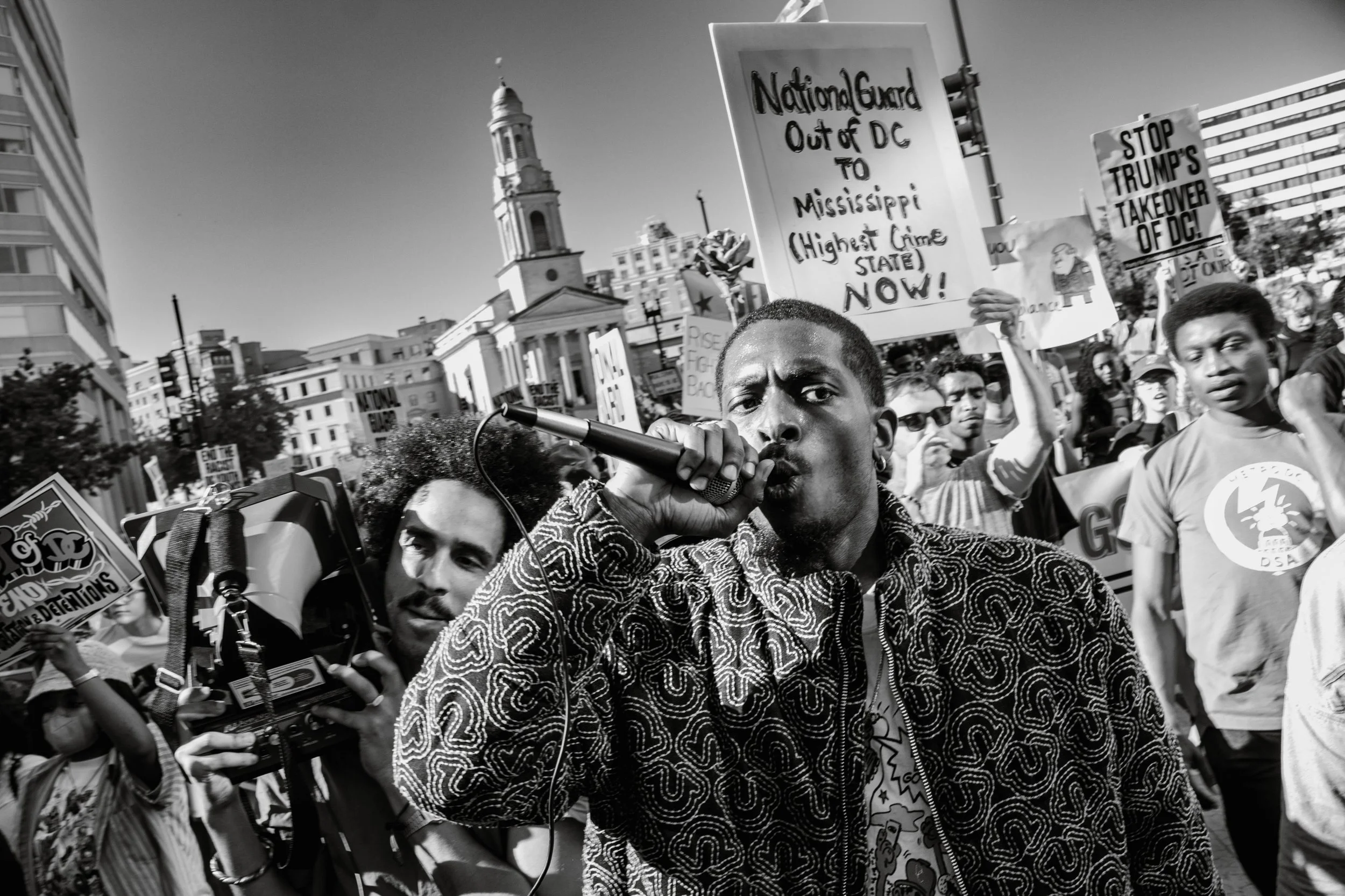 A group of protesters holding signs during a demonstration, with a man speaking into a microphone at the forefront, and a cityscape background including a church steeple.