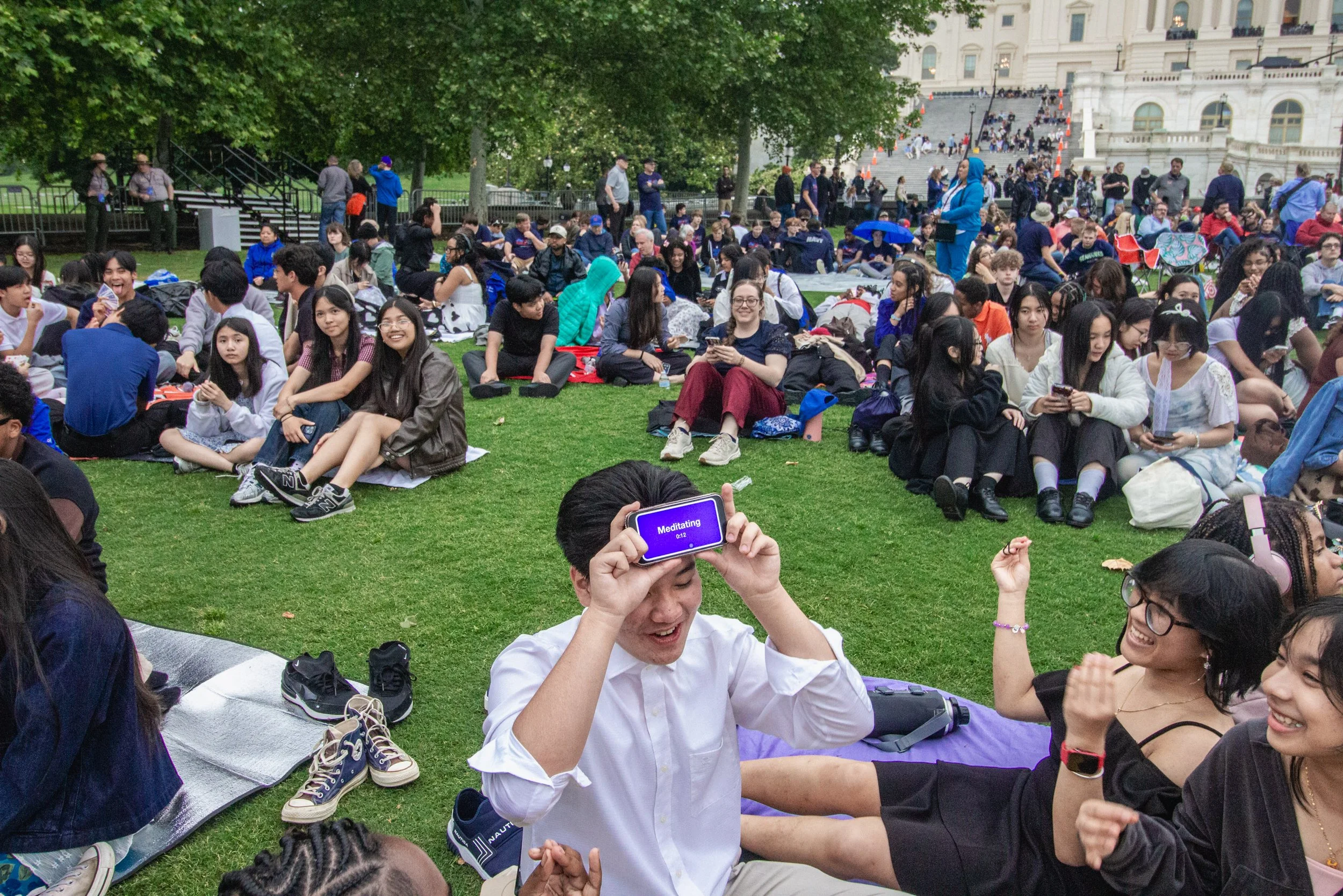 A large group of people gathered outdoors in front of a government building, some sitting on the grass, others standing around, with some participating in a session like meditation. Many are smiling and interacting, and a few are using their phones.