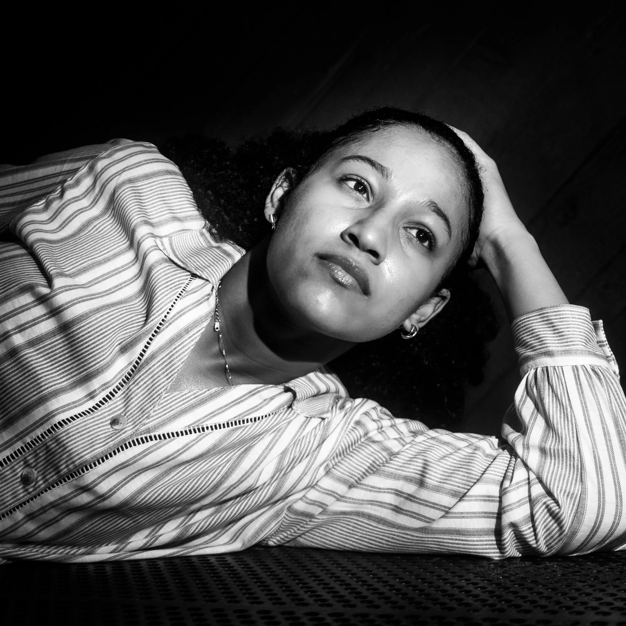 A woman lying down with her head resting on her hand, wearing a striped shirt and earrings, looking at the camera in black and white.