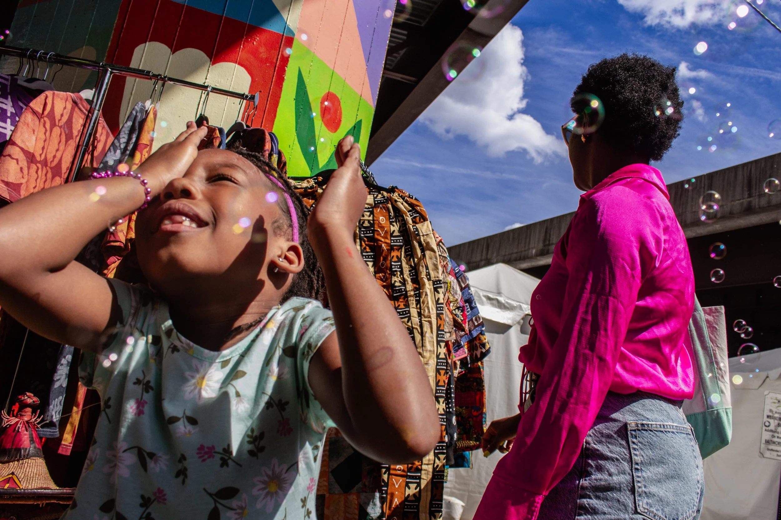 Children at an outdoor market or fair, with one girl in a floral shirt smiling and another girl with curly hair in a pink top. Colorful clothing hangs on a rack in front of a vibrant, multicolored mural. The sky is partly cloudy with lens flare and b
