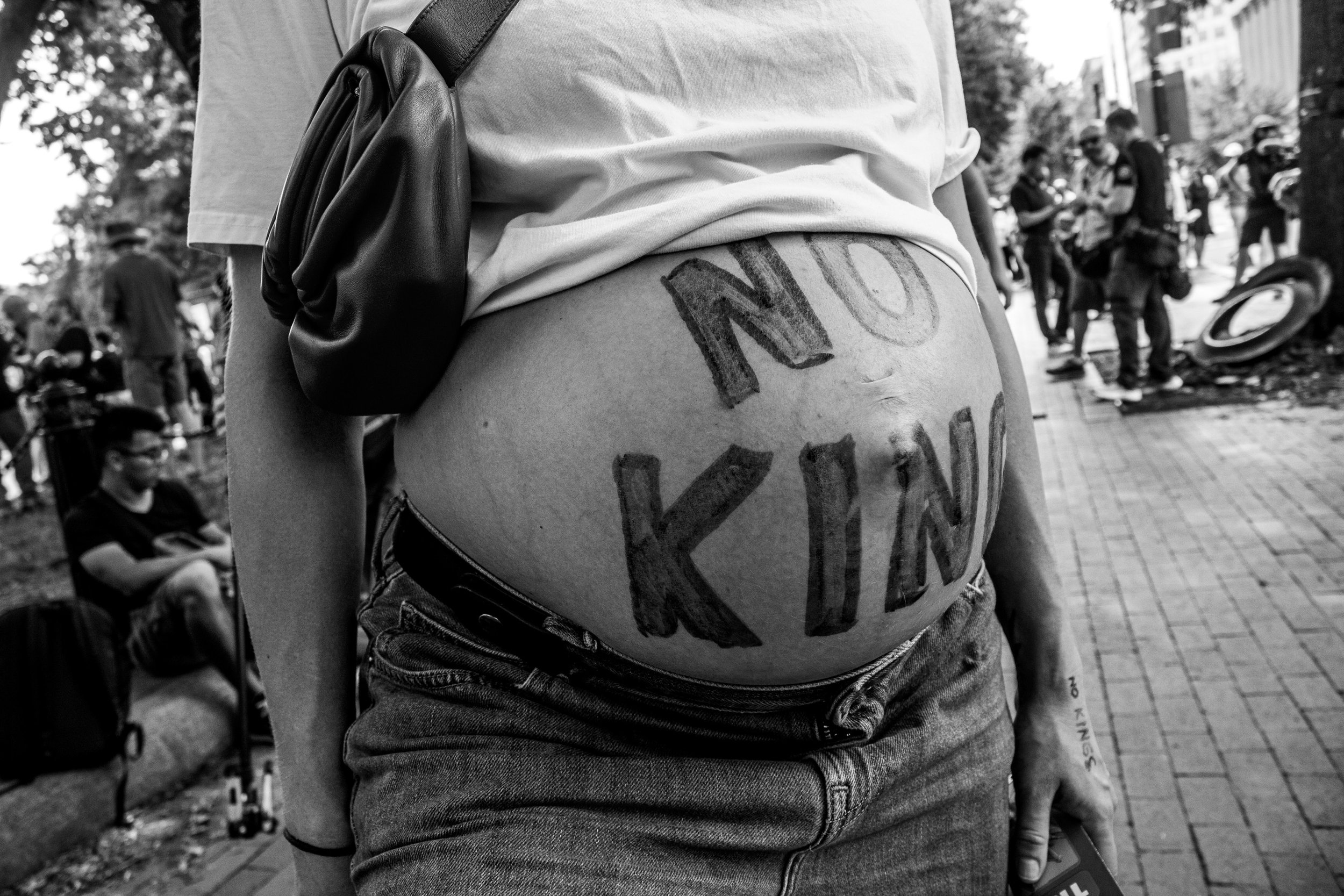 Close-up of a person's midsection with painted text on their stomach, at an outdoor gathering with several people in the background.