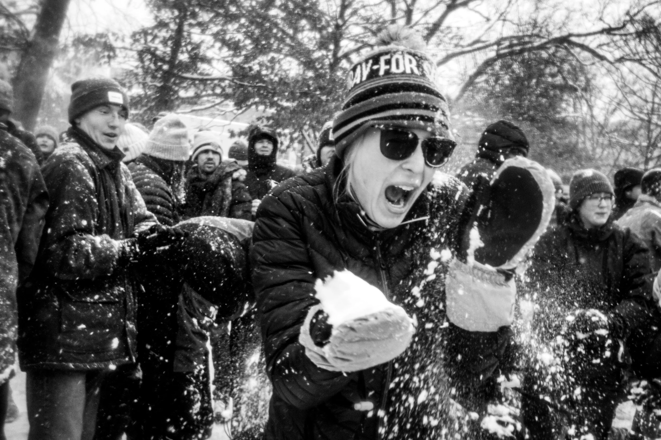 A group of people dressed in winter clothing gathered outdoors in a snowy park, with some throwing snow and one person in the foreground wearing sunglasses and a beanie, smiling and enjoying snow play.