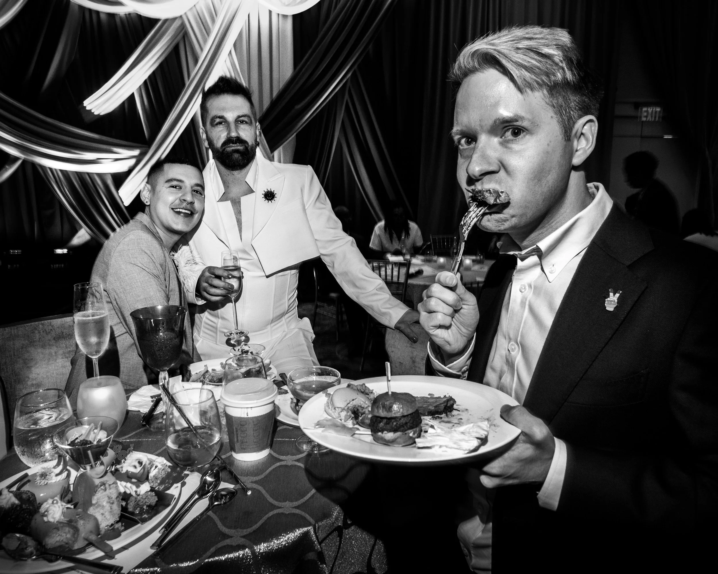 Three men at a party with food and drinks, one eating with a fork, one holding a drink, and one smiling at the camera, in a decorated venue.