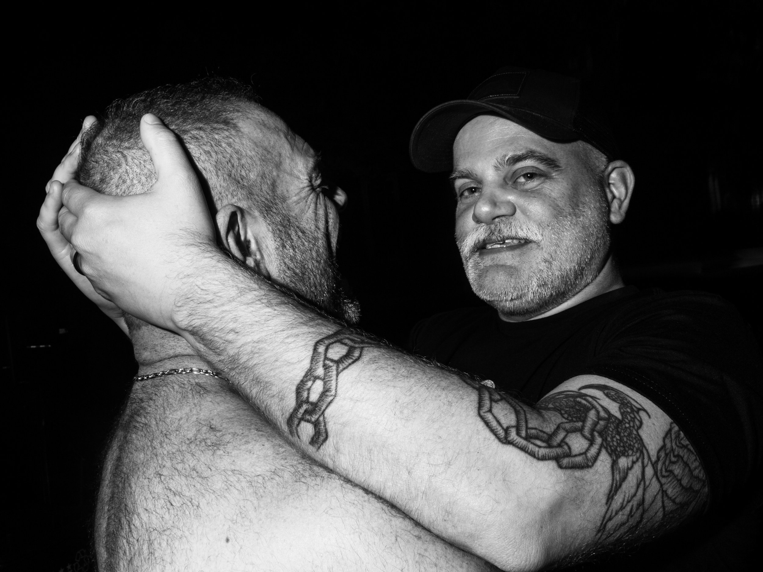 Two men, one with a cap and gray beard, the other with closed eyes, holding each other's heads closely in a kiss.