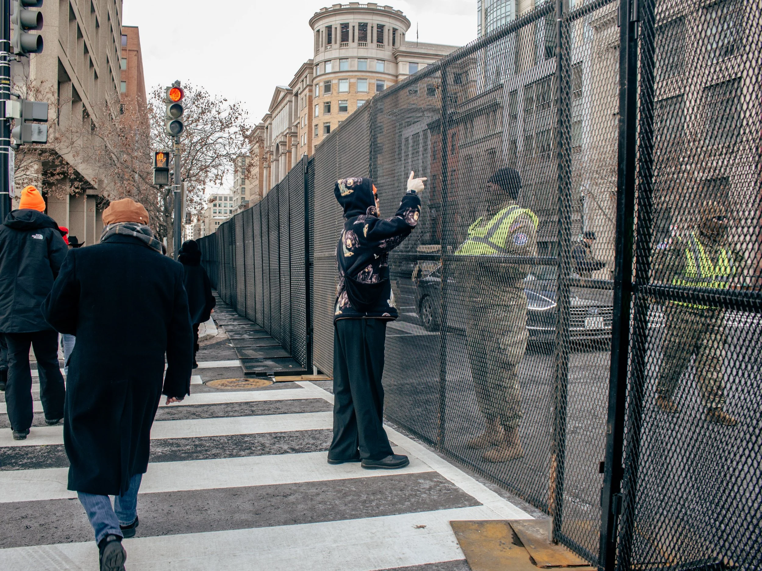 People walking on a city street near a chain-link fence with police officers inside it. A person in a black hoodie with graffiti-style designs is pointing at the officers. The background features tall buildings and a traffic signal showing a red ligh