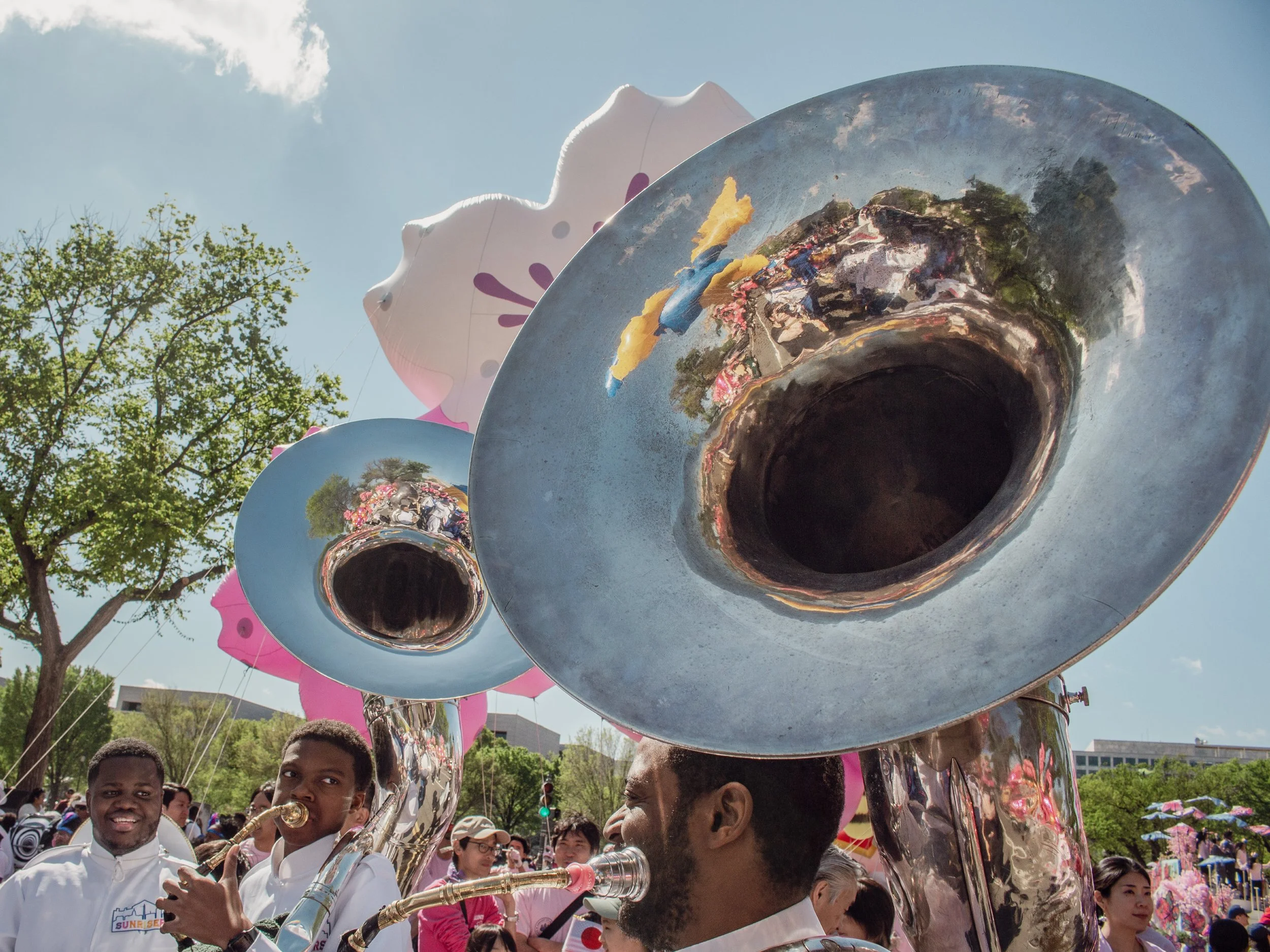 A large crowd of people outdoors, playing musical instruments including sousaphones, with balloons and trees in the background.