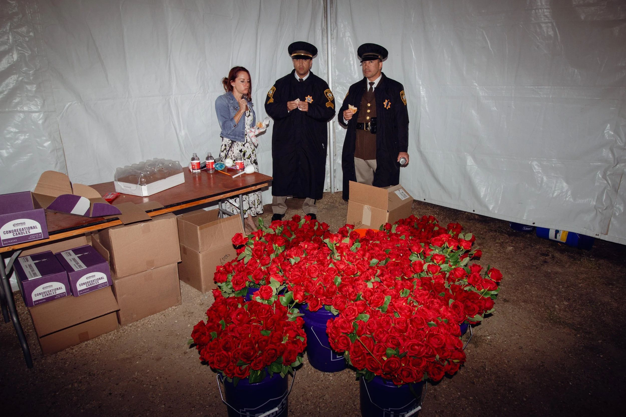 A woman and two police officers standing inside a tent, eating food. The table next to them has boxes of purple candles, bottles of water, and other small items. In front of the table, there are large buckets filled with red roses. The space appears 