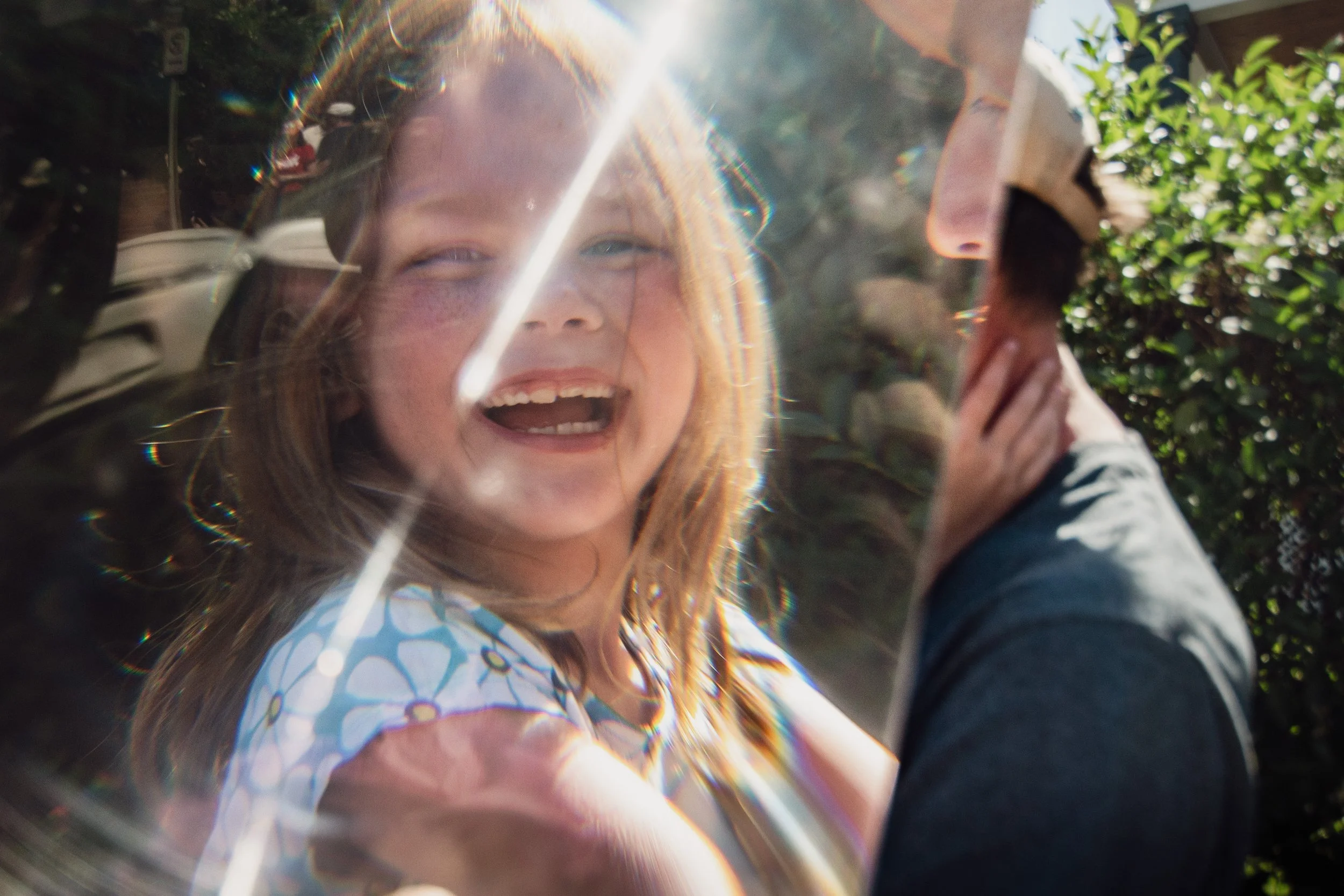 A young girl with reddish hair and freckles smiling and waving in front of a camera, with sunlight creating lens flare and rainbow-like optical effects. There is greenery, a street, and a person partially visible on the right side of the image.