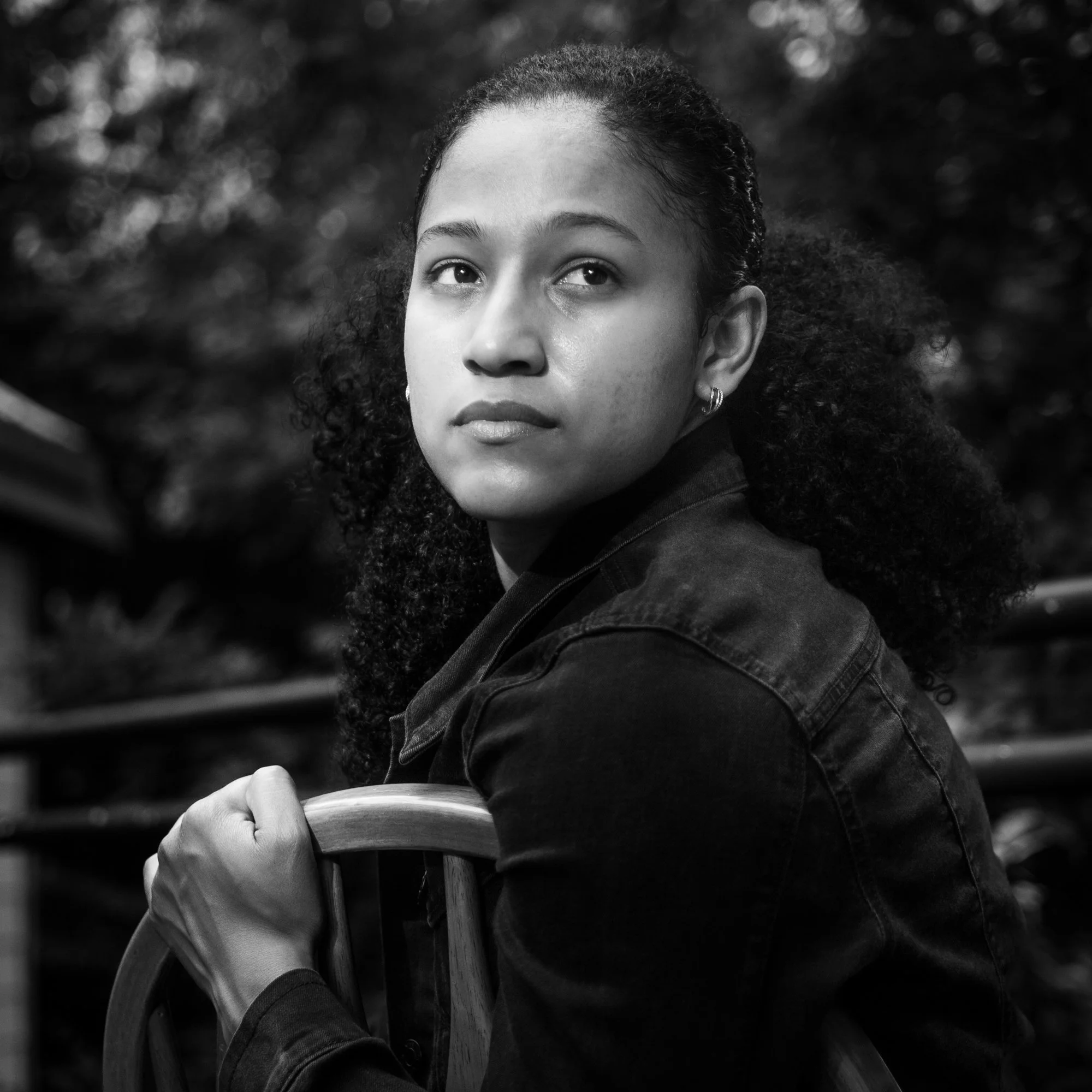 A young woman with curly hair looking slightly to the side and upward in an outdoor setting, with blurred trees in the background, in black and white.