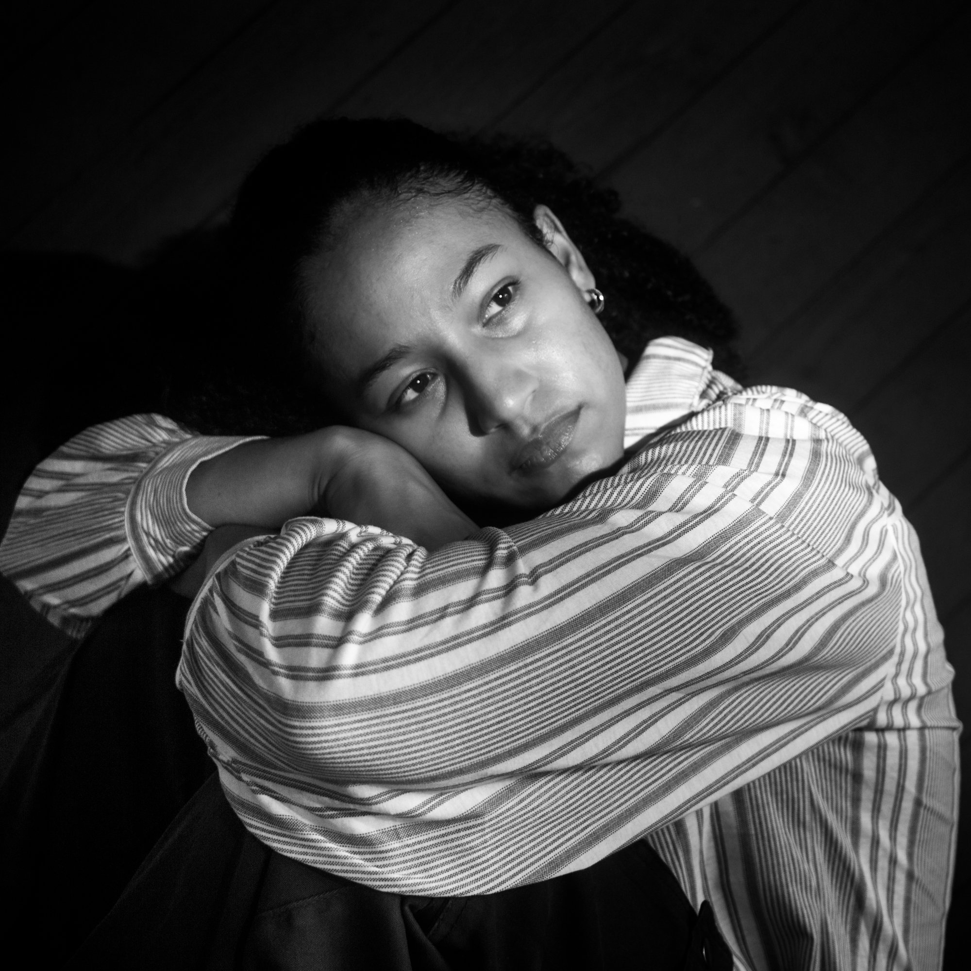 Close-up of a woman lying down on a dark surface, smiling softly with her head resting on her arm. She has curly hair, earrings, and is wearing a striped shirt.