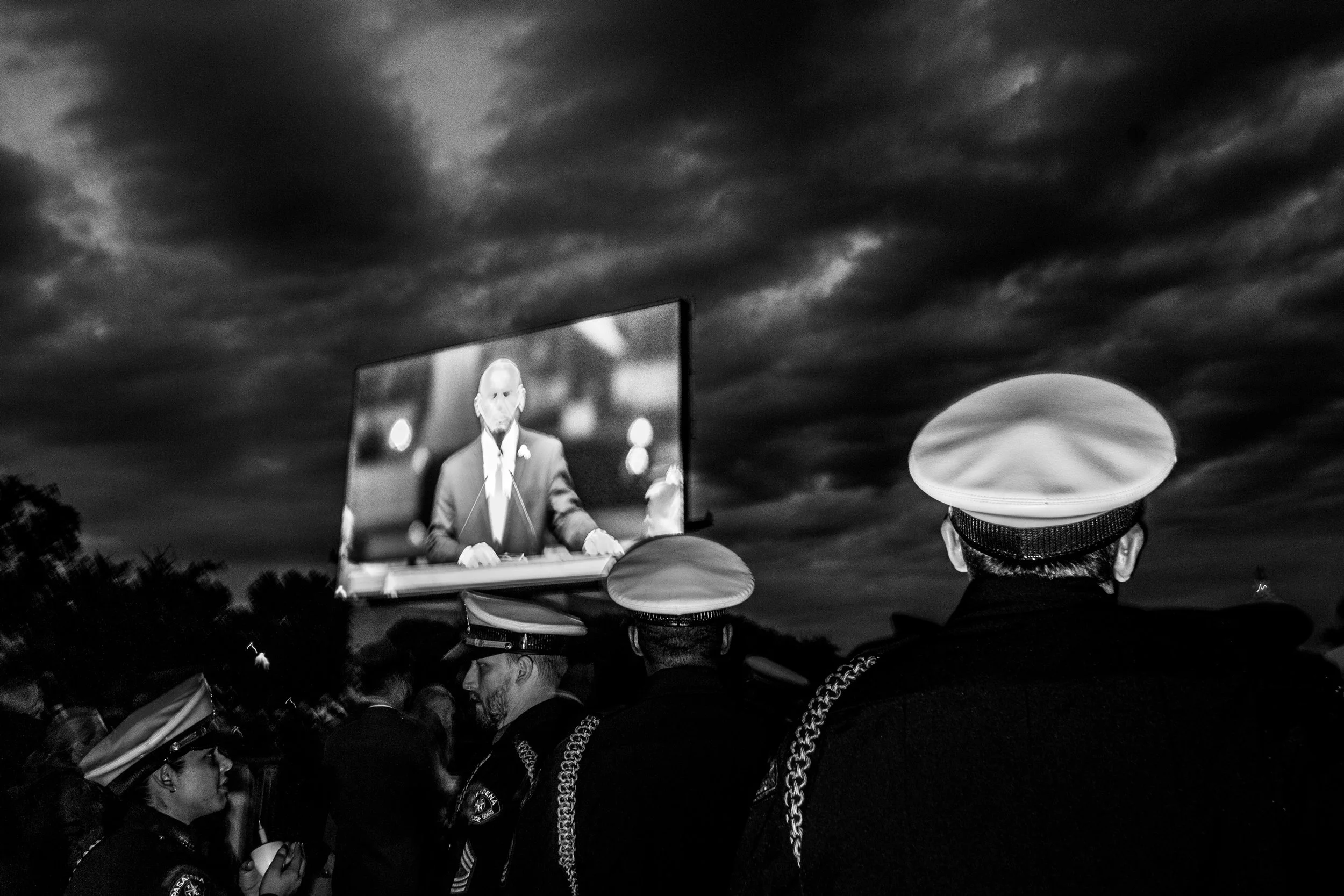 A crowd of people, some wearing chains and police uniforms, gather outdoors on a cloudy evening. In the background, a large screen displays a man in a suit speaking at a podium.