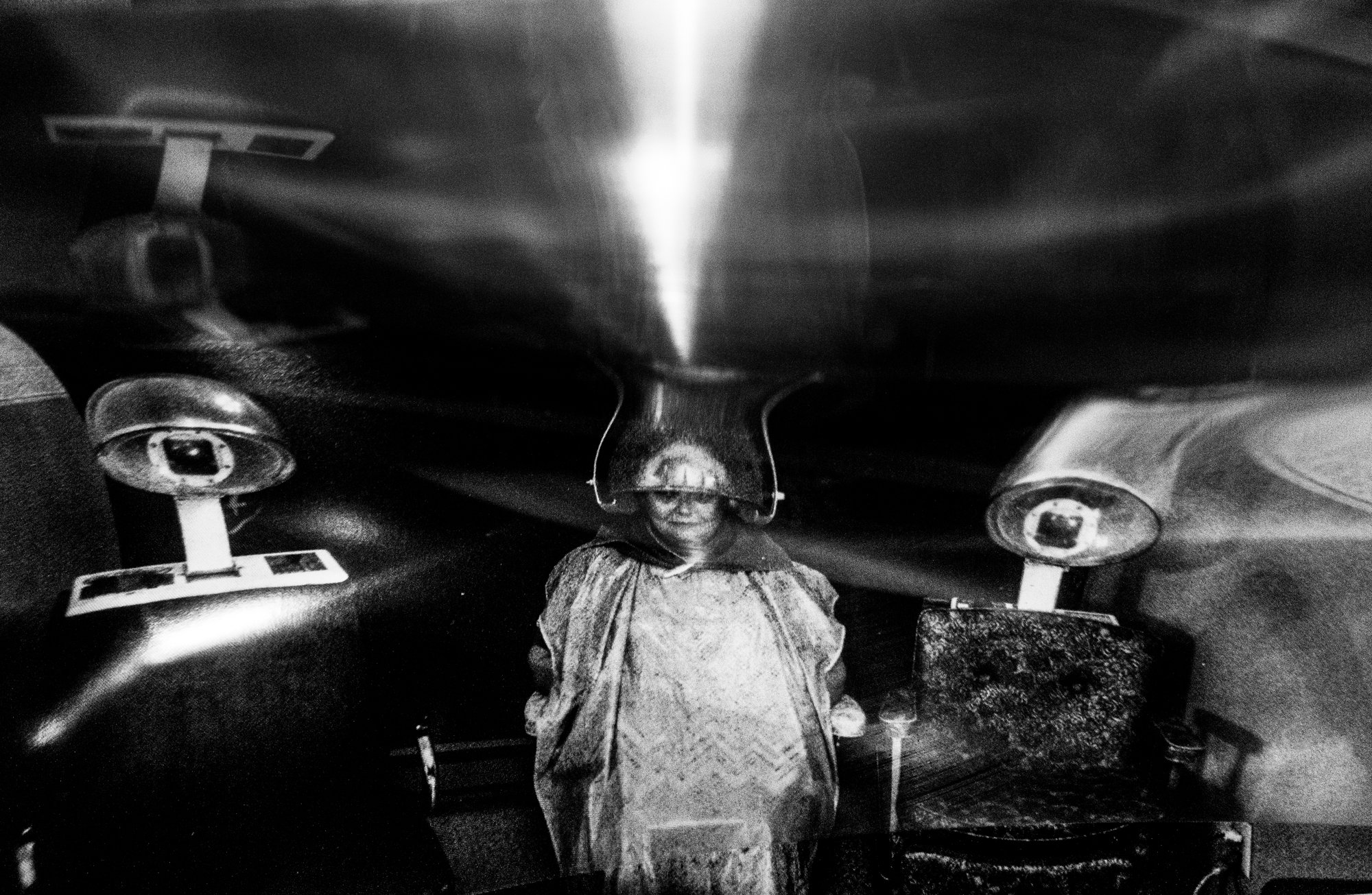 A black and white photo of a doll with a circular face, framed by a hat, placed on a reflective surface with ceiling lights overhead.