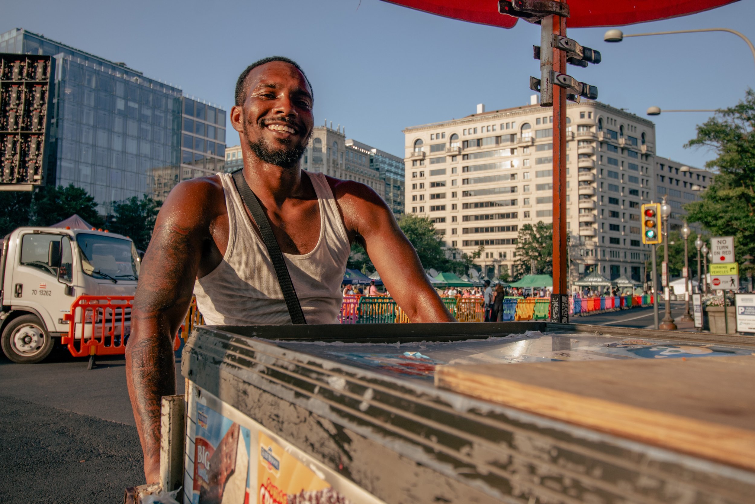 Smiling man with short dark hair and beard, wearing a sleeveless shirt, standing behind a food cart in an urban setting with tall buildings, streetlights, and a traffic light in daylight.