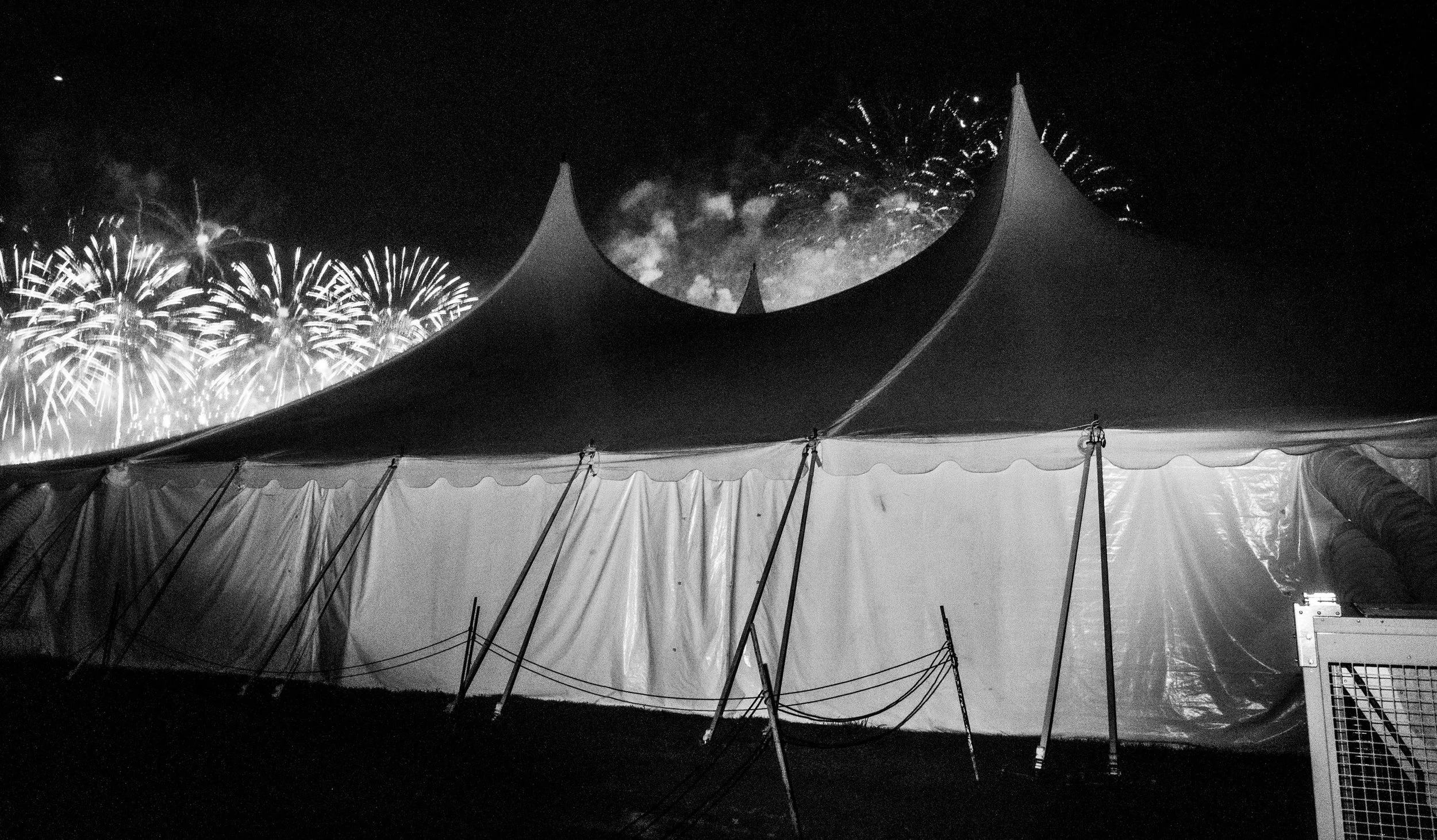 Nighttime fireworks display above a circus tent with pointed peaks, viewed from inside the tent with support poles and fabric visible in the foreground.