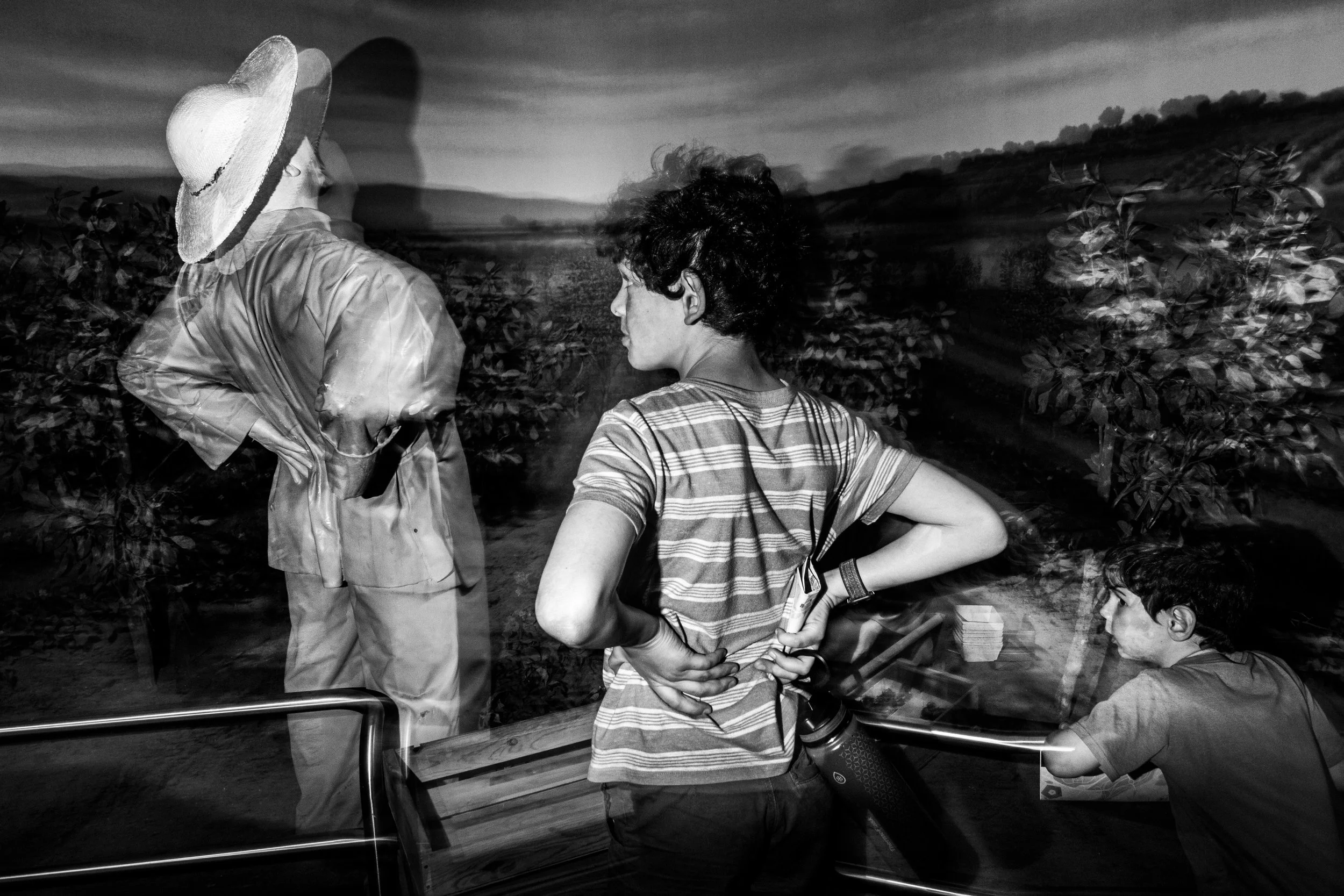 Black and white photo of a woman and a young girl on a boat ride looking at a scenic landscape with greenery and mountains in the distance. The woman is wearing a wide-brimmed hat and a raincoat, while the girl is dressed in a striped shirt and holds