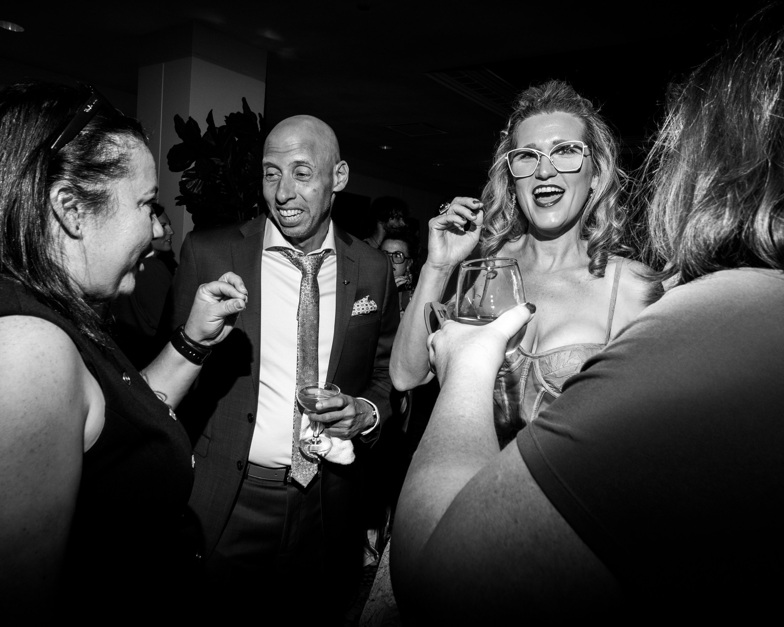 Four people at a social event, two women and two men, engaging in conversation and holding drinks. One woman with glasses is smiling and sticking out her tongue, while the other woman on the left is smiling and facing the group. The man in the middle