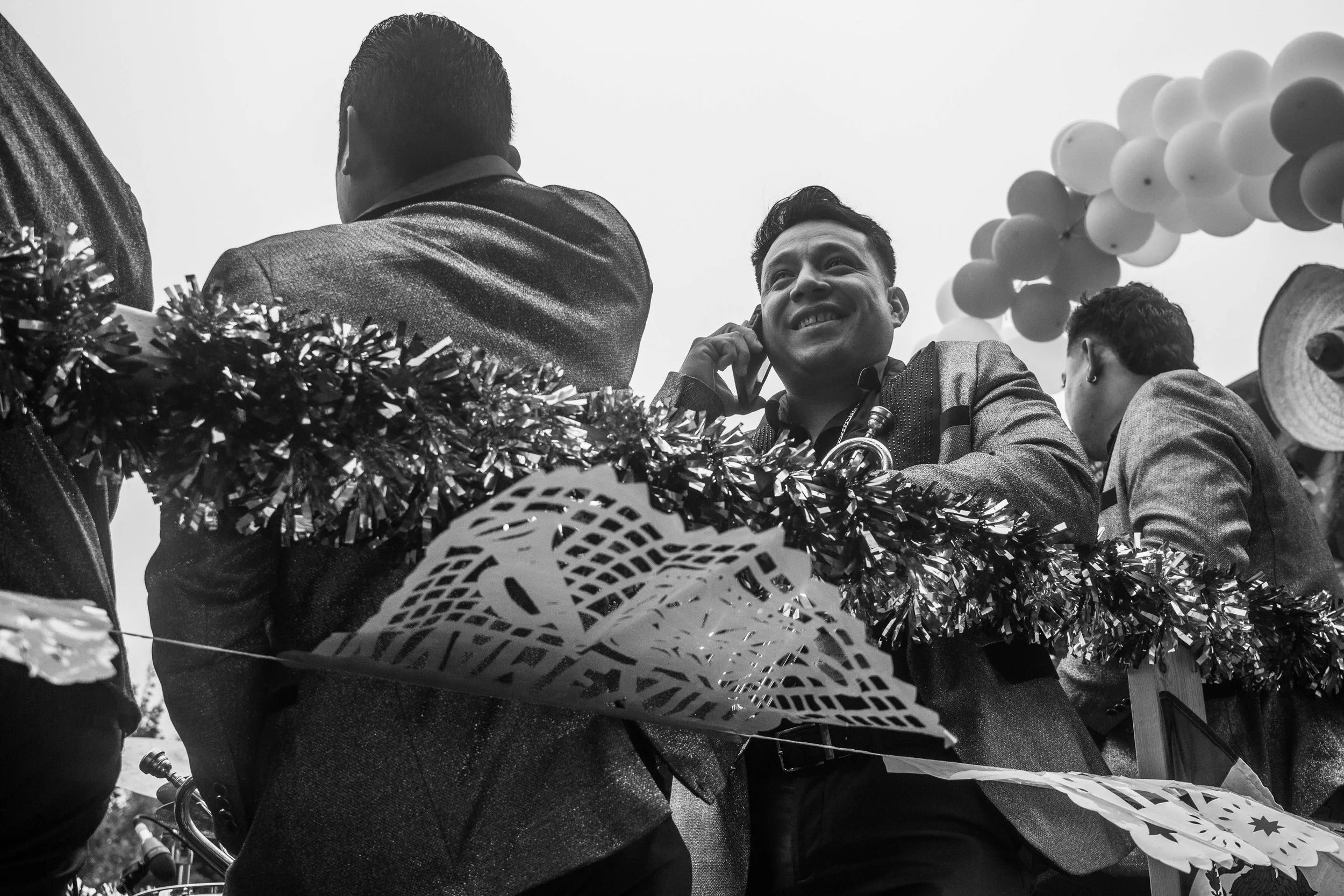 Men dressed in formal suits at a celebration event, decorated with balloons, tinsel, and paper cutouts, with one man smiling and talking on a phone.