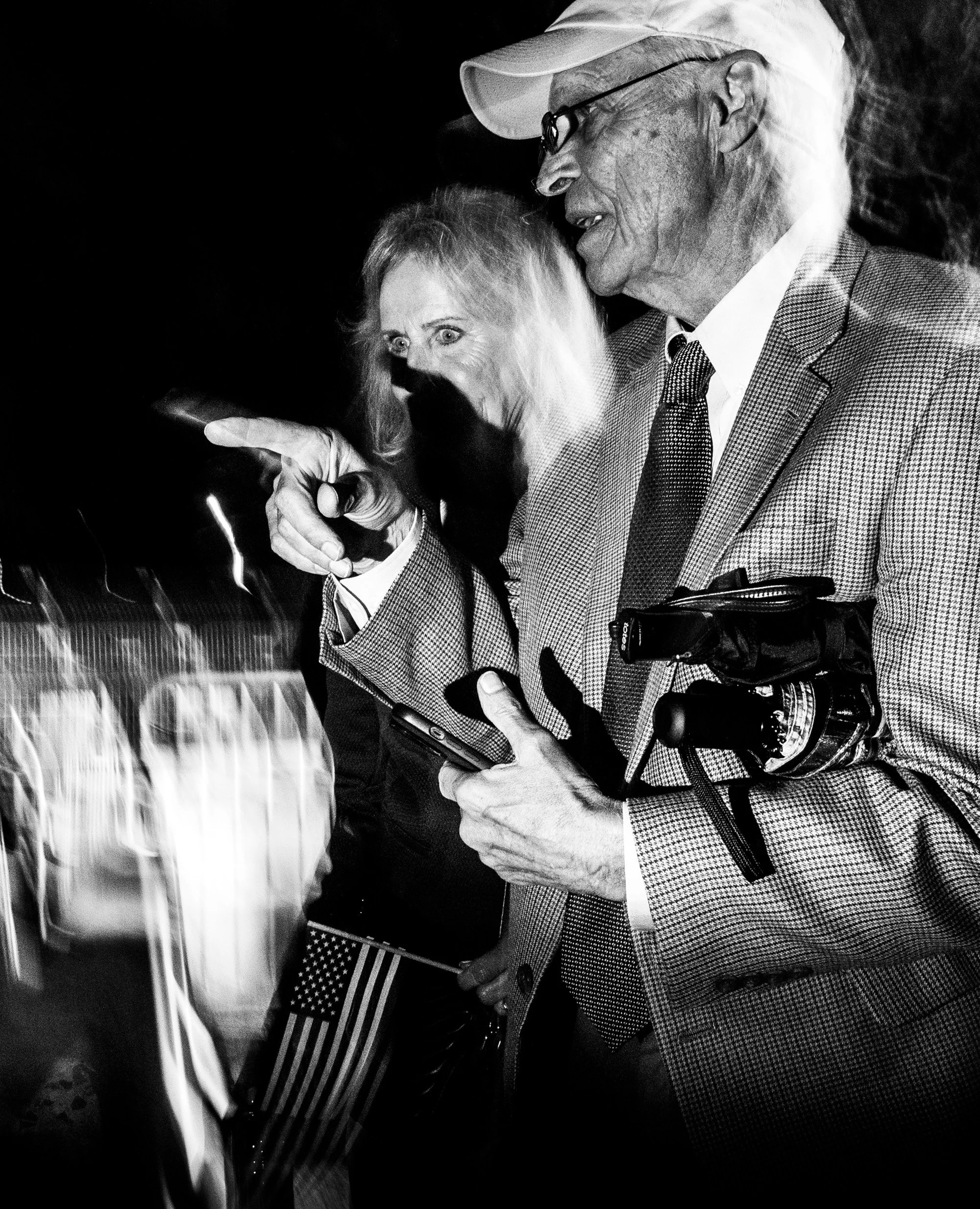 An elderly man with glasses wearing a light-colored hat, checkered blazer, and dress shirt, holding a smartphone in his right hand, appears to be smiling or talking to someone. A woman with light hair, wearing a dark top, is standing close to him, po