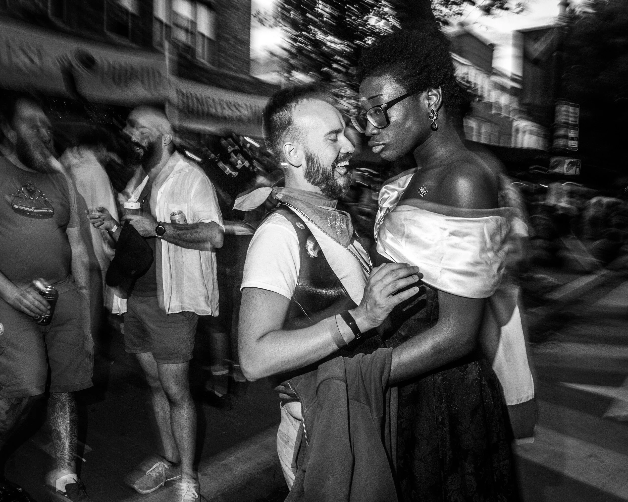 A black and white photo of a couple dancing closely in a crowded street at night, with people and storefronts blurred in the background.