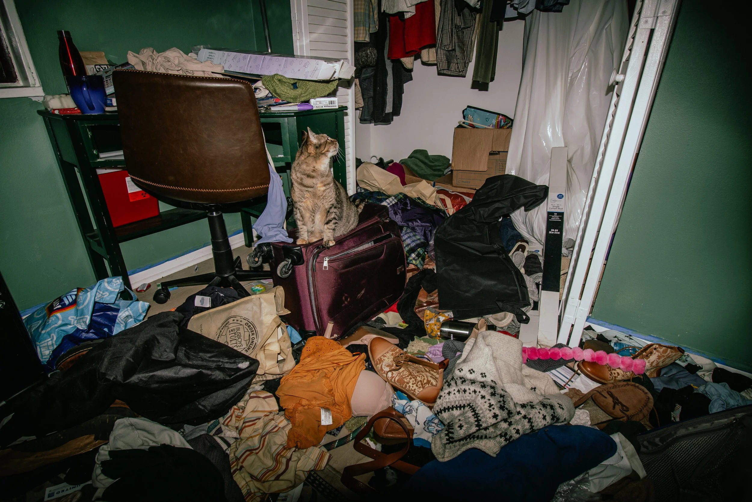 A cluttered room with a brown office chair, an open closet with clothes hanging, and various clothes and items scattered on the floor. A tabby cat is sitting on a maroon suitcase in the center of the room.