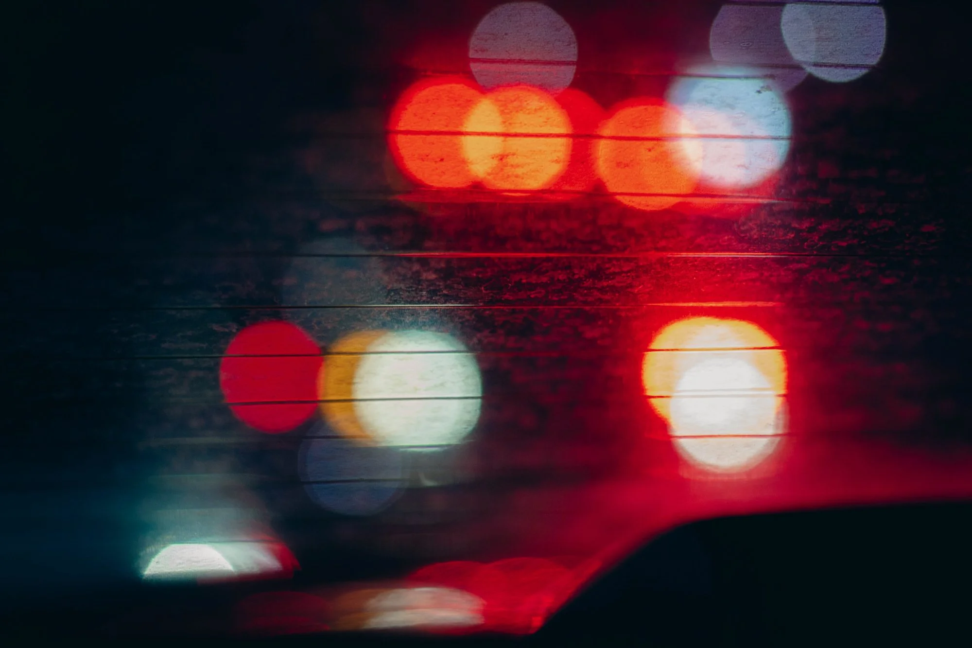 Blurred car brake lights seen through a window with raindrops at night, creating a bokeh effect.