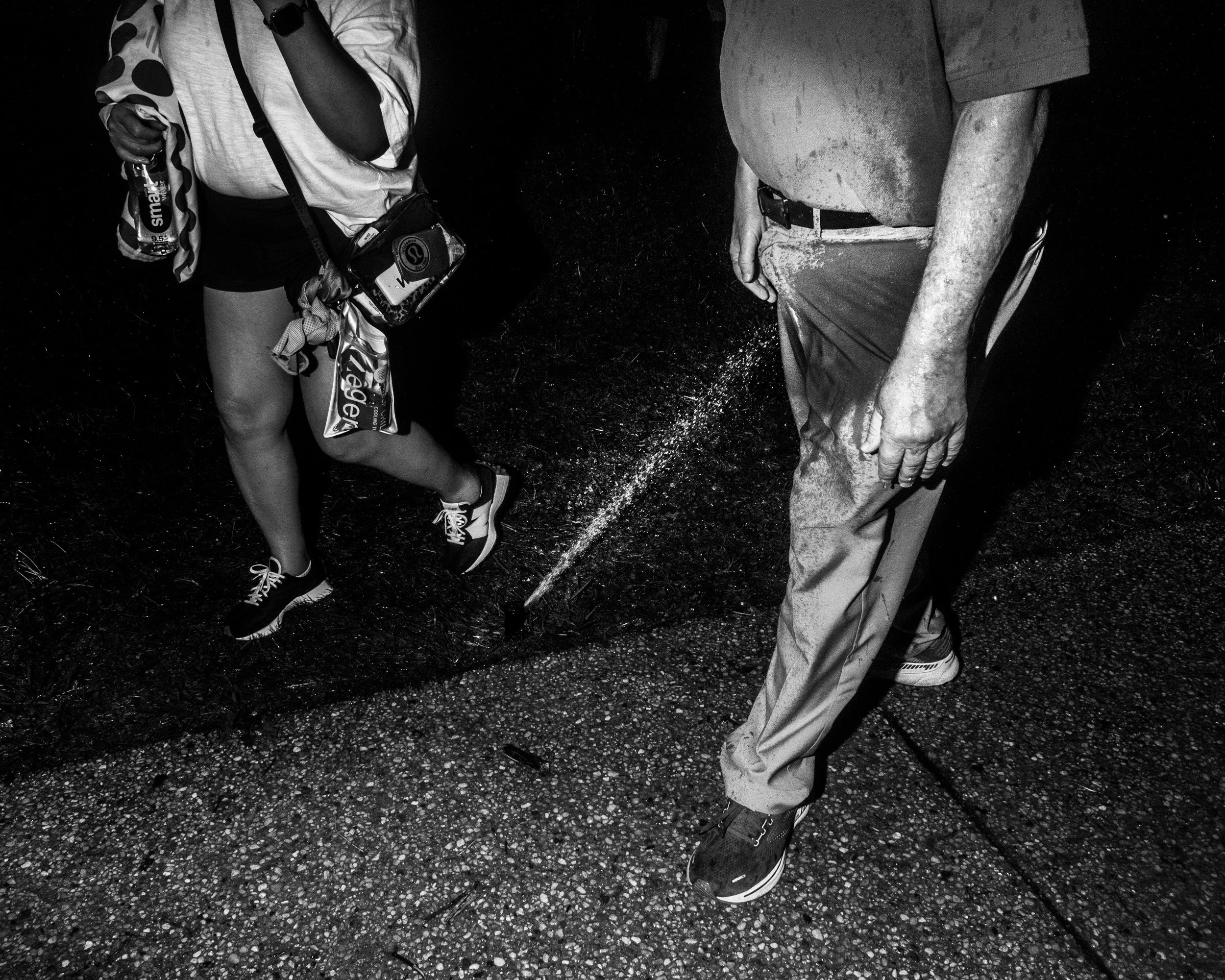 Close-up of two people standing on a gravel surface, one holding a water bottle, the other with hands clasped. Both are wearing casual clothes, and the scene appears to be outdoors at night.