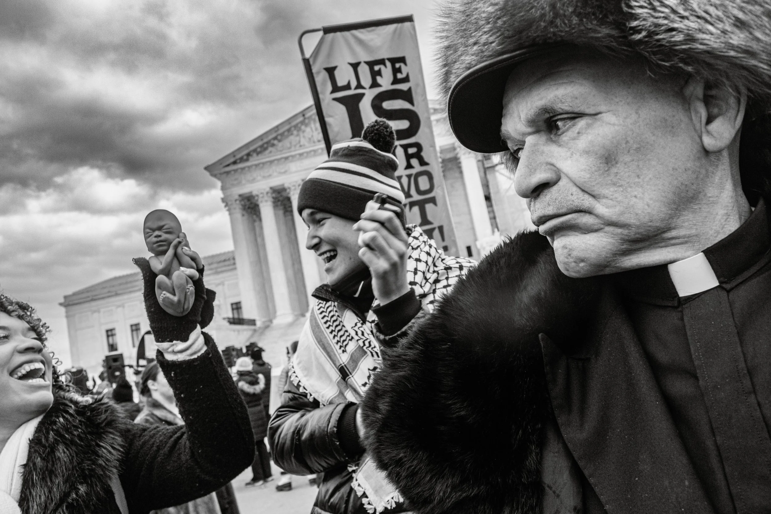 Black and white photo of a group of children and adults participating in a public demonstration in front of a government building. One child holds a puppet, and a person holds a sign that says 'LIFE IS' with the rest of the message obscured. The grou