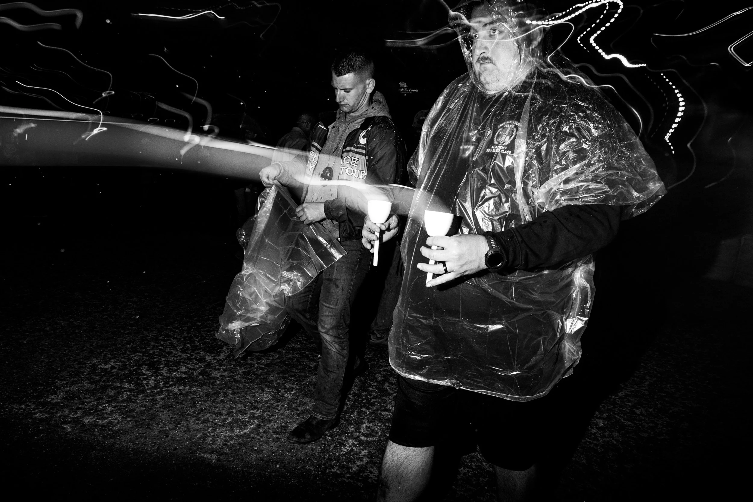 Two men at night, one in a reflective rain poncho holding two cups, the other looking at a phone or device, with blurred light streaks surrounding them.