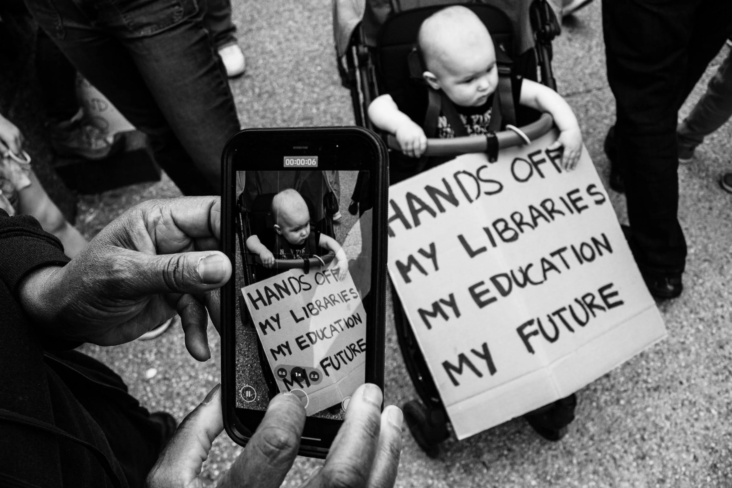 A small child with a shaved head sitting in a stroller holds a protest sign that reads 'HANDS OFF MY LIBRARIES EDUCATION FUTURE'. Someone is taking a photo of the child with a smartphone, capturing the scene.