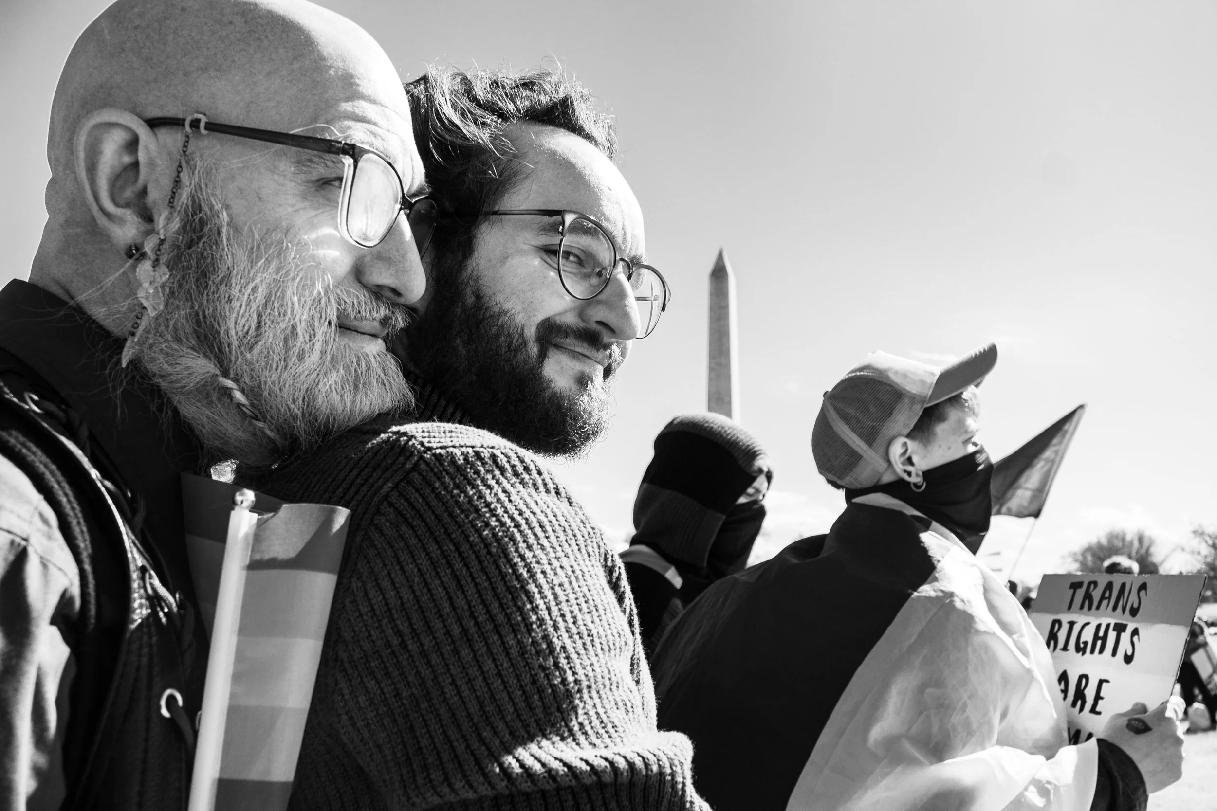 Black and white photo of three people standing outdoors, holding signs, with the Washington Monument visible in the background. They are wearing glasses and warm clothing, participating in a demonstration or rally.