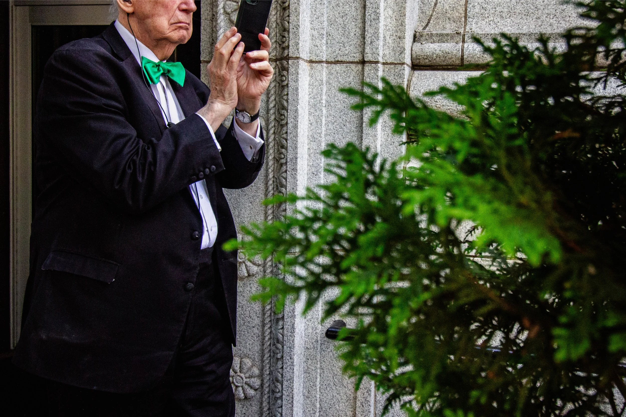An elderly man in a black suit with a green bow tie is standing outdoors near a stone building, holding a phone to his ear, with greenery in the foreground.