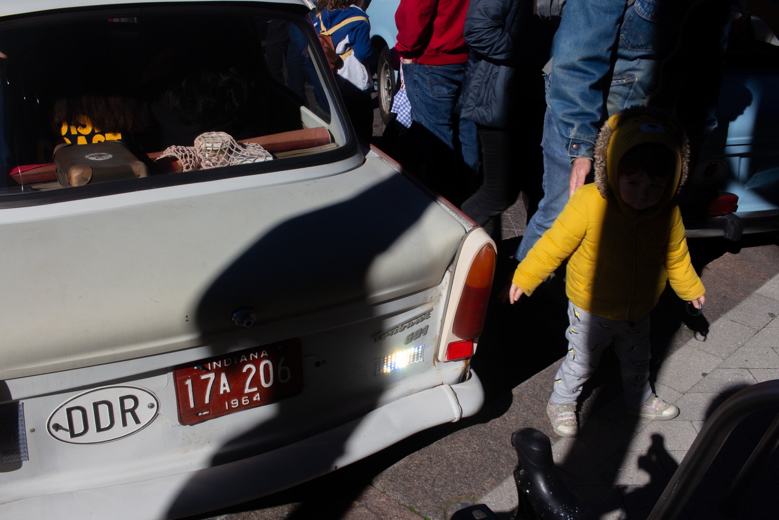 Close-up of the back of a small white vintage car with a deer head sticker and red Indiana license plates, surrounded by people and a child in a yellow jacket.