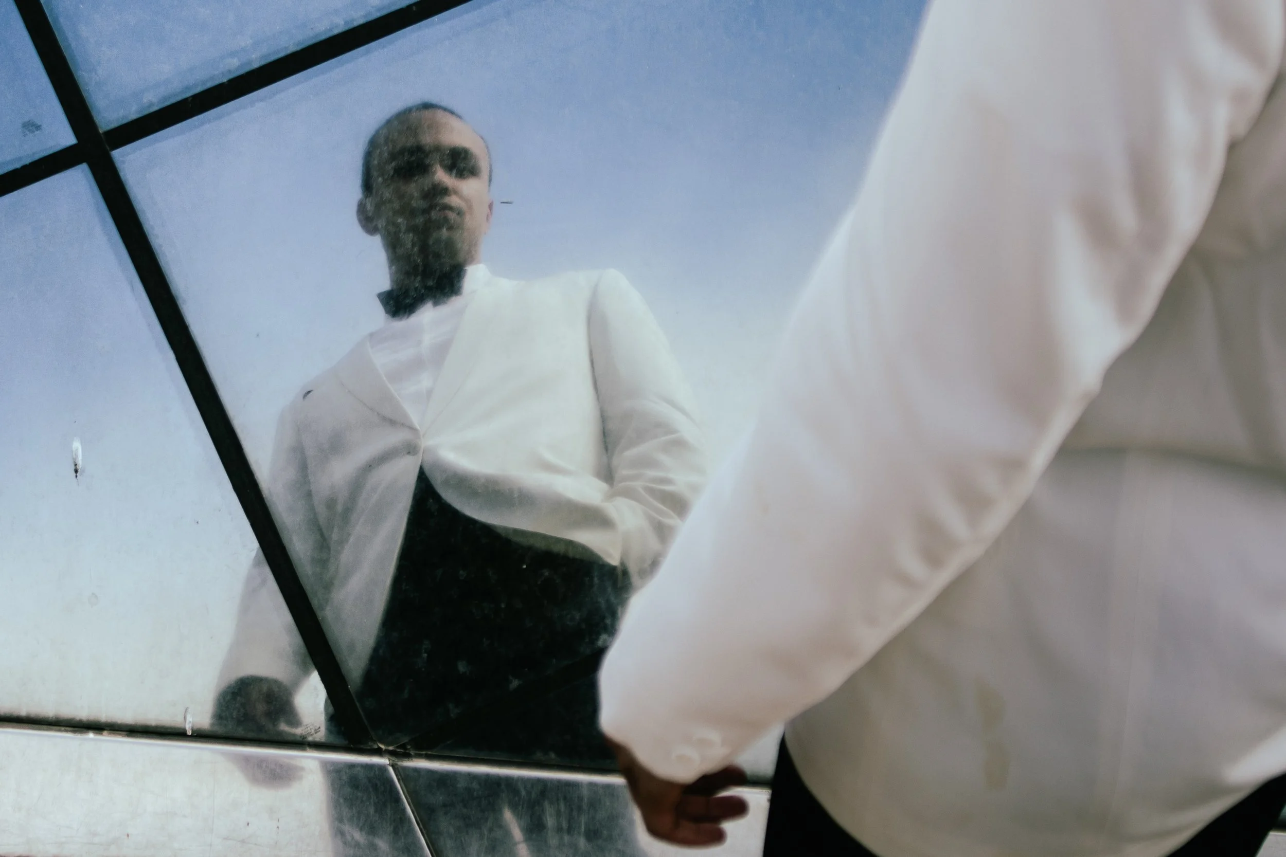 Reflection of a man in a mirror, wearing a white jacket, with a blue sky and grid lines visible in the background.