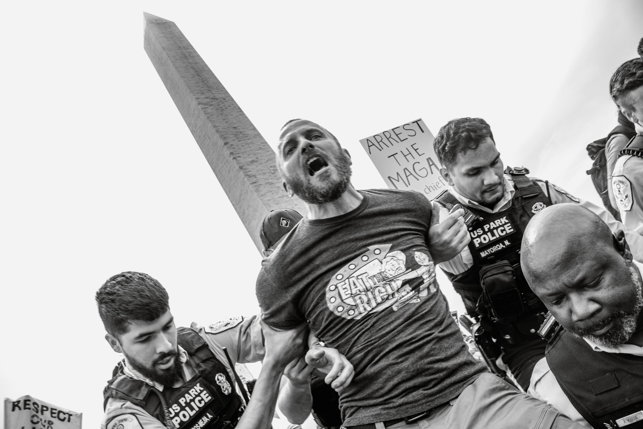 A black and white photo of protestors, including police officers and a man shouting, holding a sign that says "ARREST THE MAGA" with some people wearing police uniforms and others with signs that say "RESPECT".
