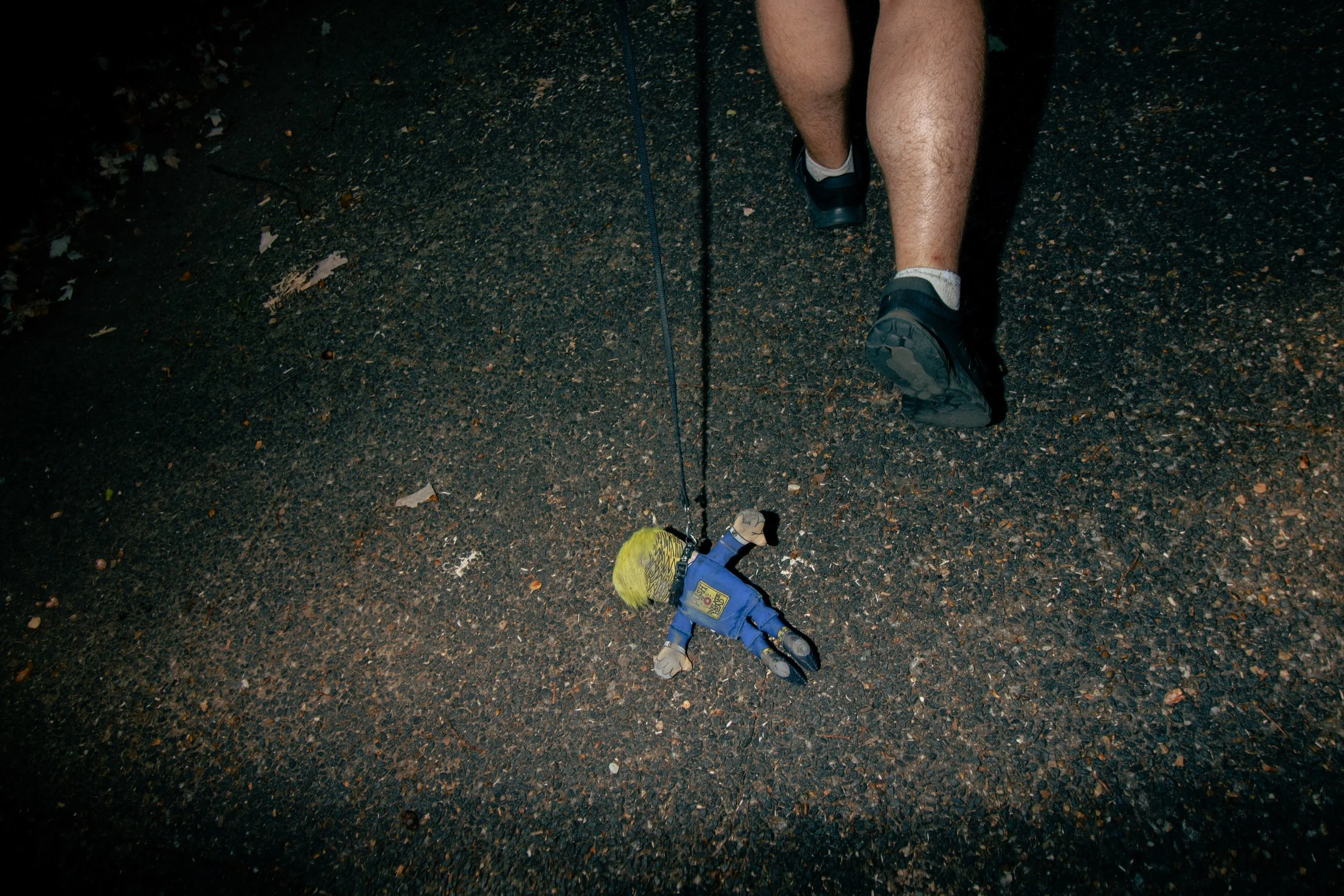 Nighttime outdoor scene of a person walking on a wet, dark asphalt path with fallen leaves, wearing black shoes, gray socks, and beige shorts. A small, colorful toy dog on a blue leash is on the ground.