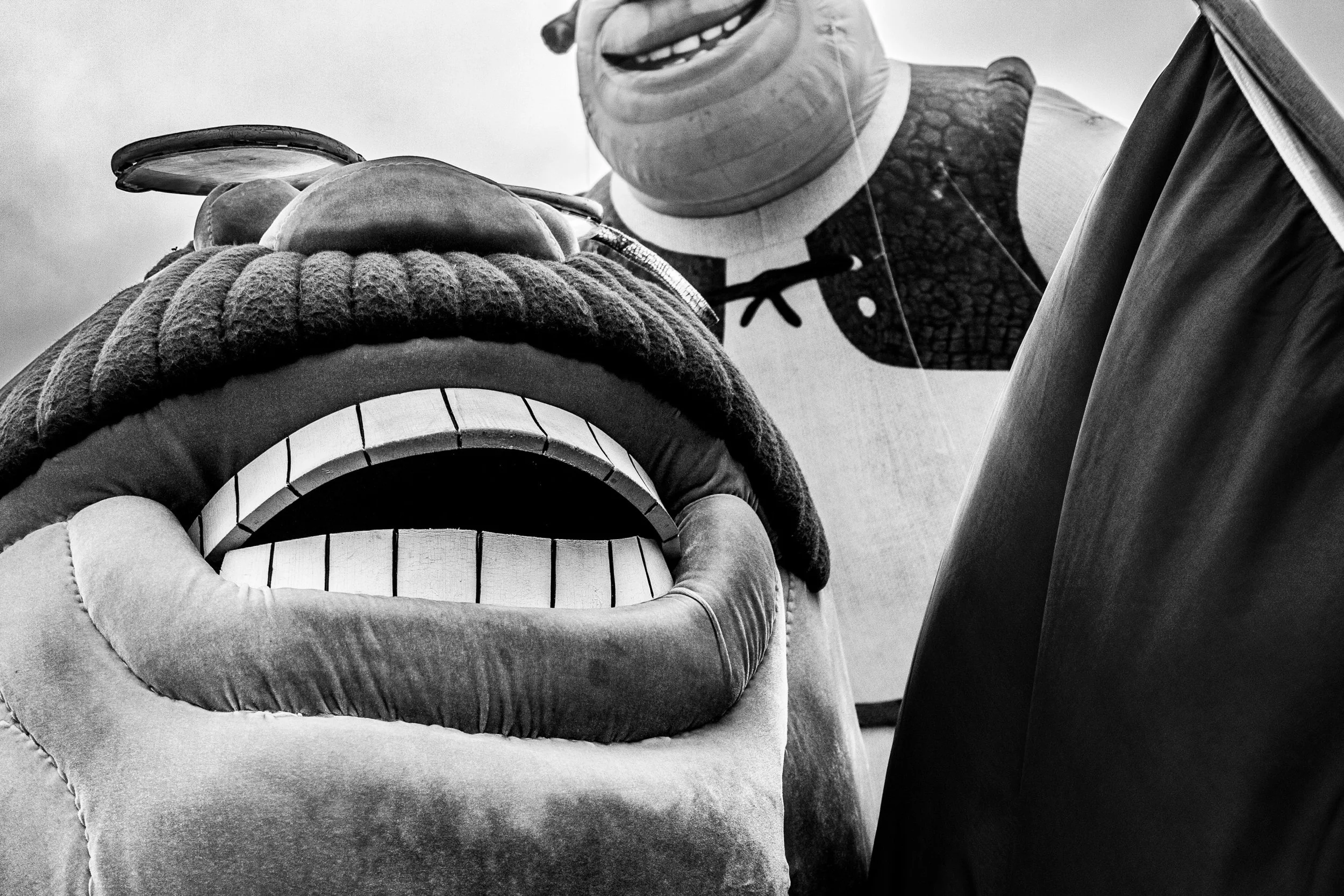 A large puppet or mascot resembling a character with a big, open mouth, large teeth, and textured skin, possibly inspired by a dragon or monster, in front of a person holding a balloon shaped like a character with a wide smile and dark textured body.